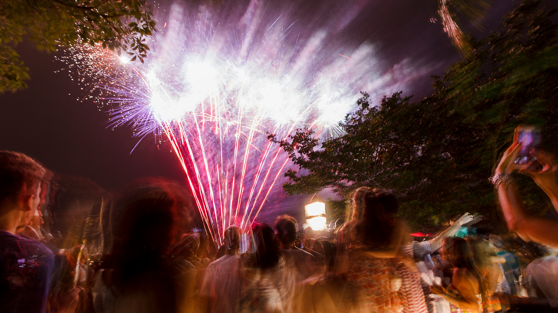 Nighttime celebration scene with fireworks lighting up the sky, and a crowd of people watching and taking photos. Some trees are visible in the surroundings.