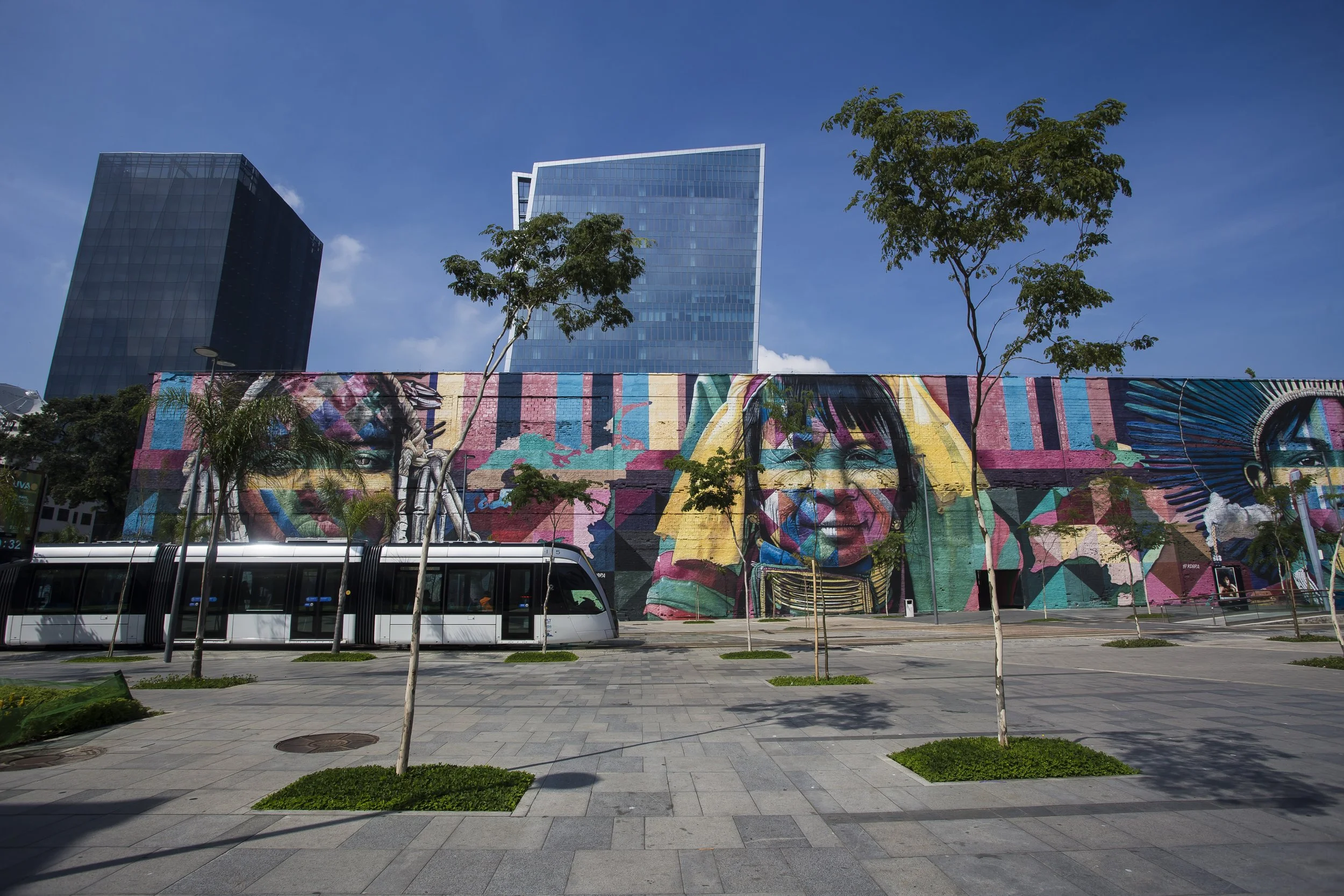 Colorful mural featuring diverse faces on a large wall in an urban area, with modern glass skyscrapers and leafless trees in the background, under a clear blue sky.
