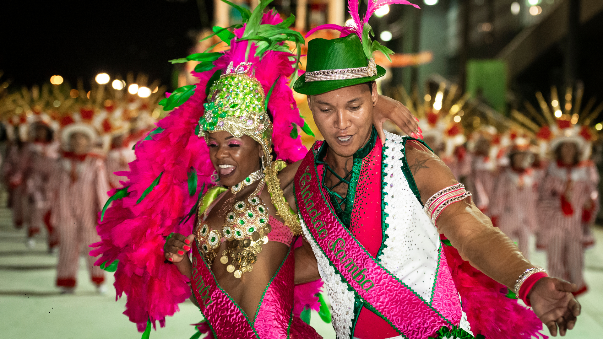 Couple dancing in colorful costumes during a parade or carnival at night, with dancers in striped outfits in the background.