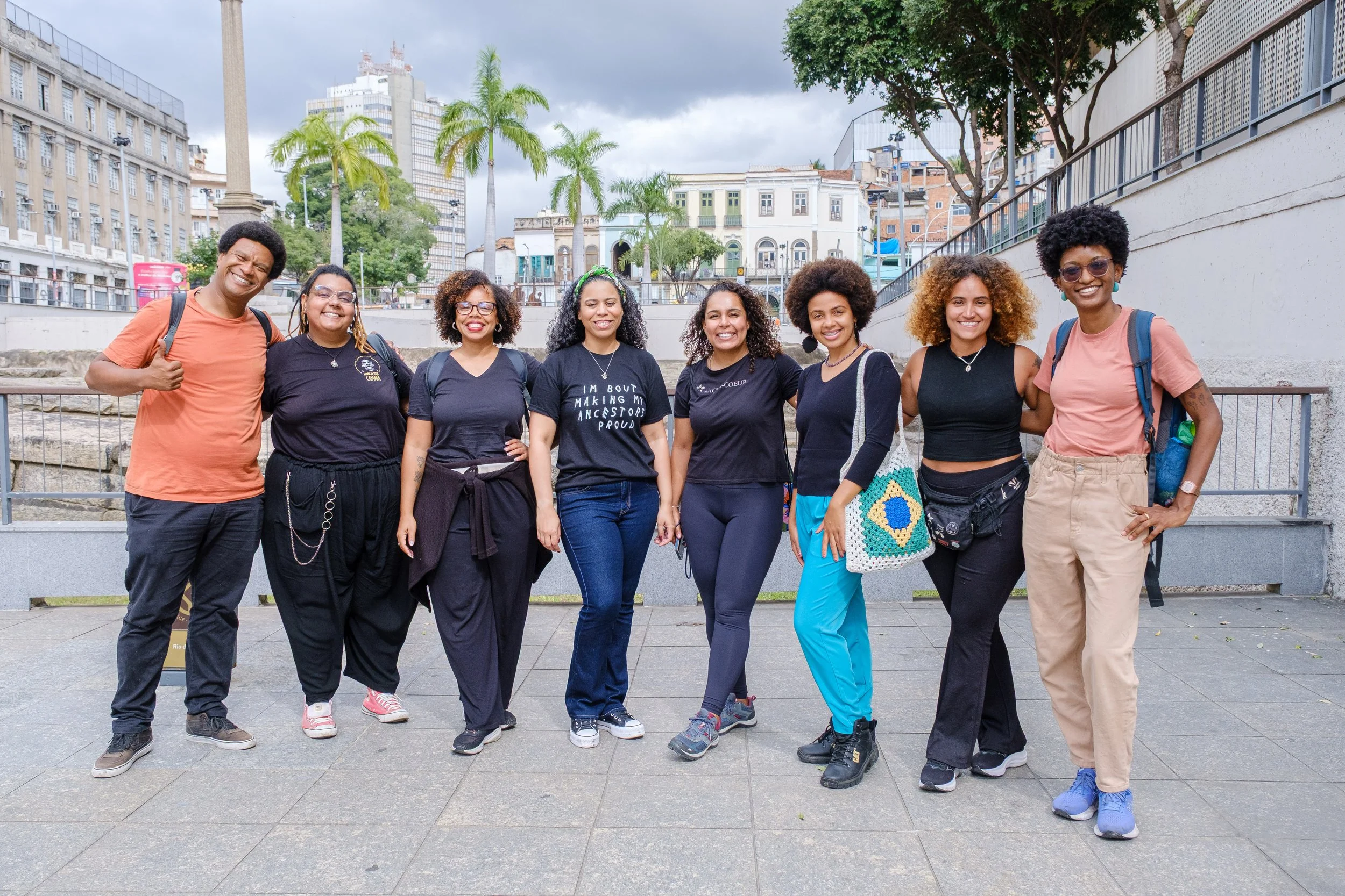 Group of diverse smiling women and one man standing outdoors in an urban park with buildings in the background, some palm trees, and cloudy sky.