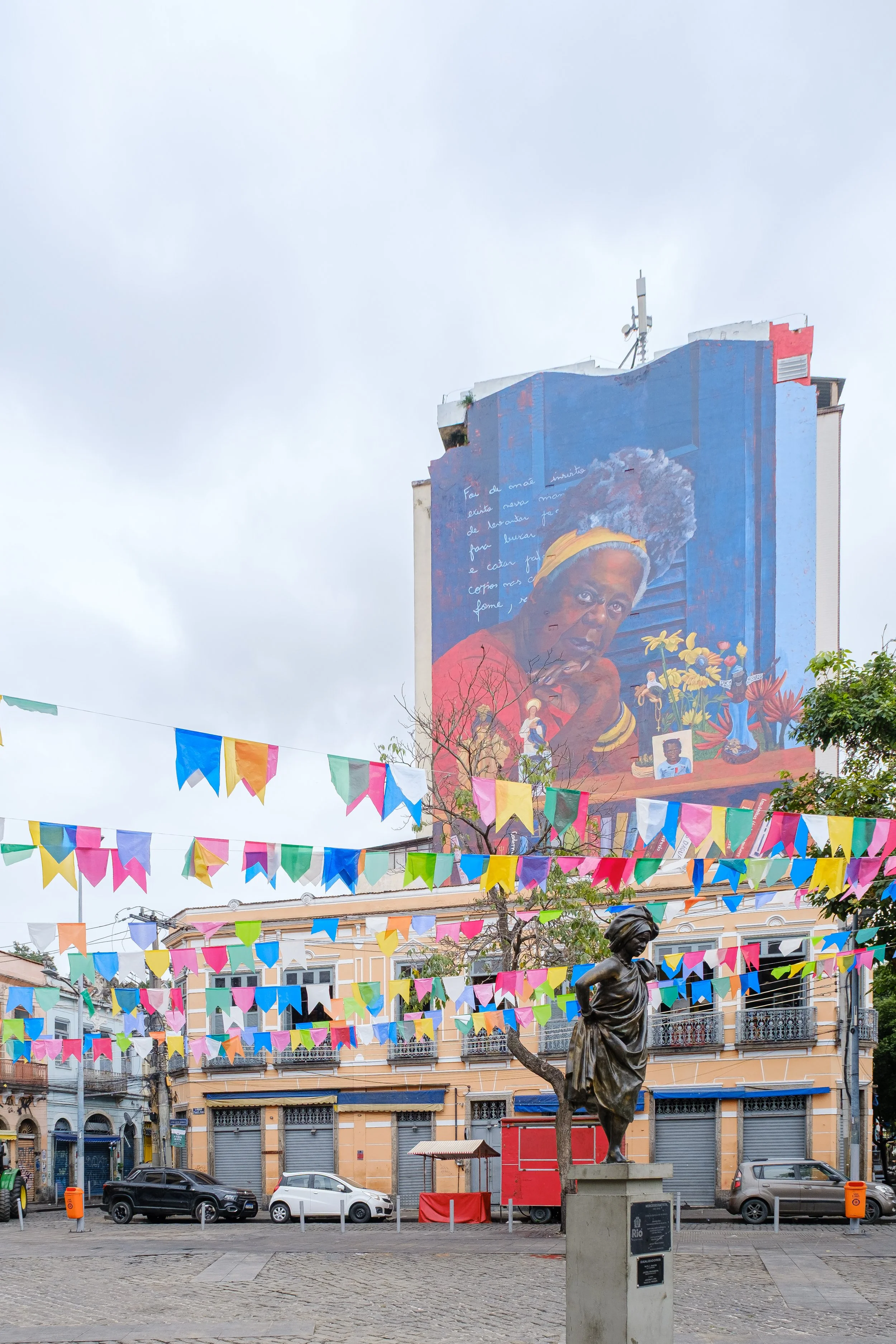A city square with cars parked along the street, colorful paper banners hanging overhead, a statue of a woman holding a branch, and a large mural of a woman with curly hair and yellow headscarf on a building. The mural contains text in a foreign language.