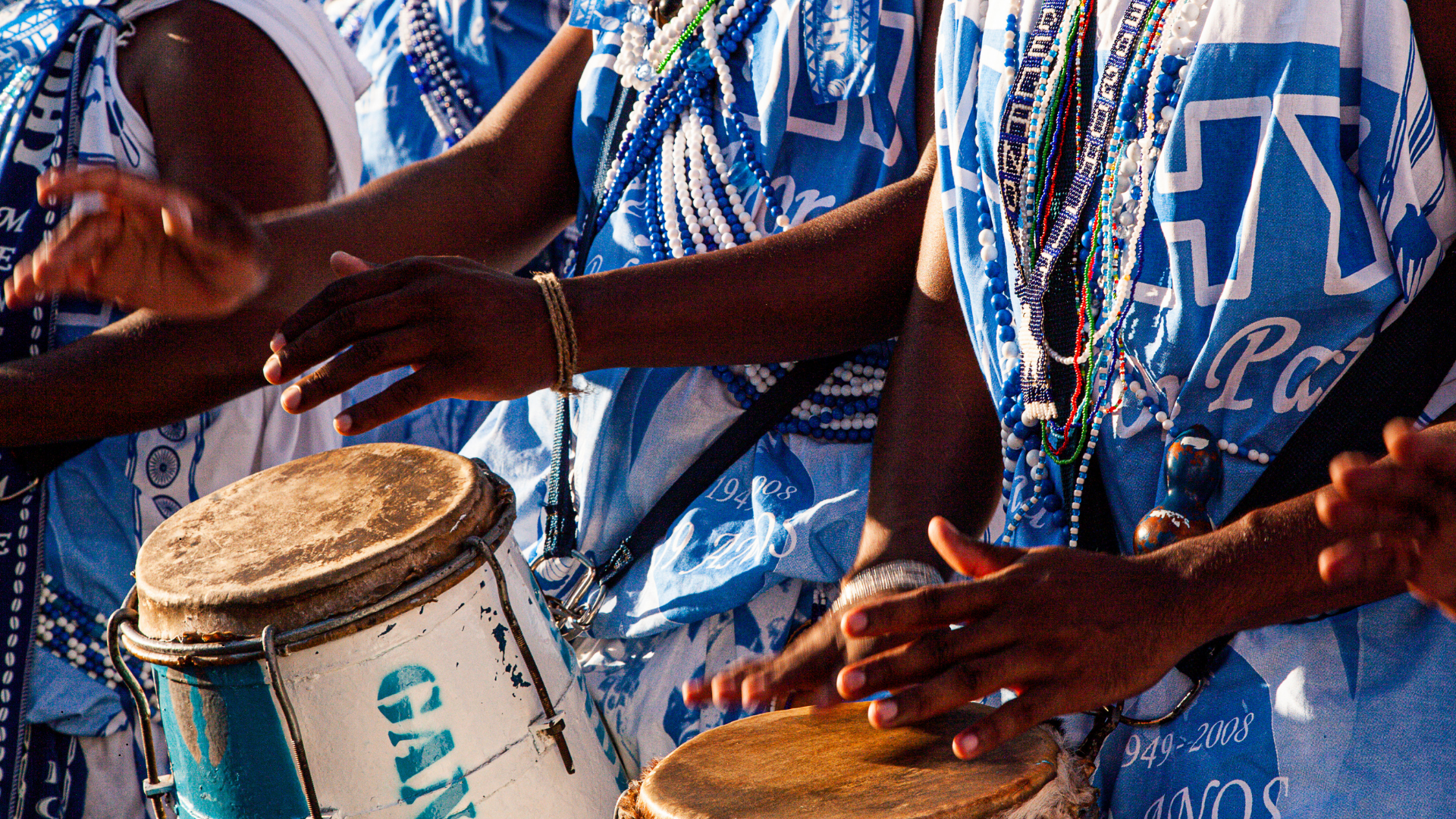 Group of people wearing blue and white traditional clothing, playing drums and wearing beaded necklaces.