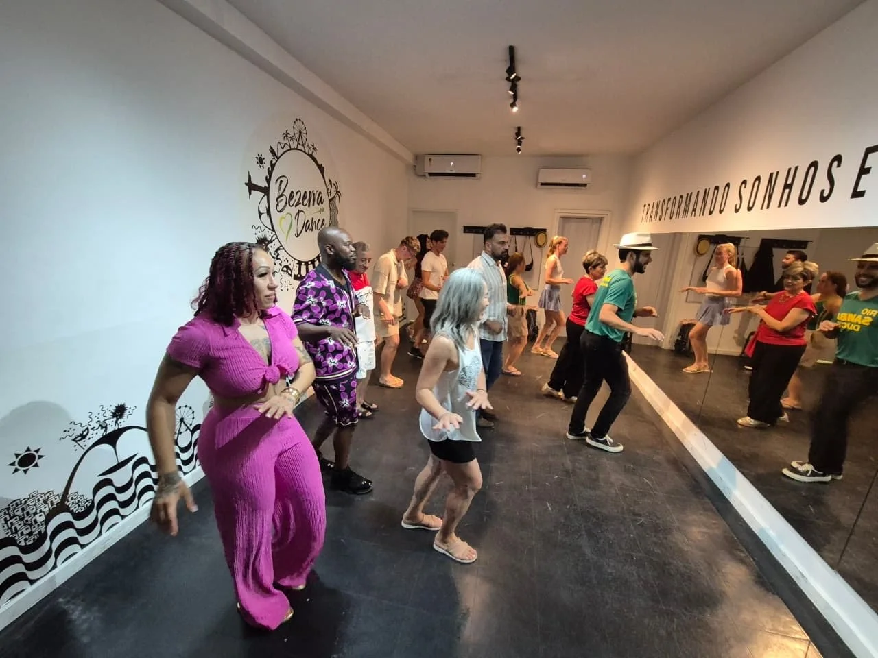 Group of people participating in a dance class in a studio, with some individuals dancing in front of a mirror and others watching, under wall art that says 'Transformando Sonhos e'.