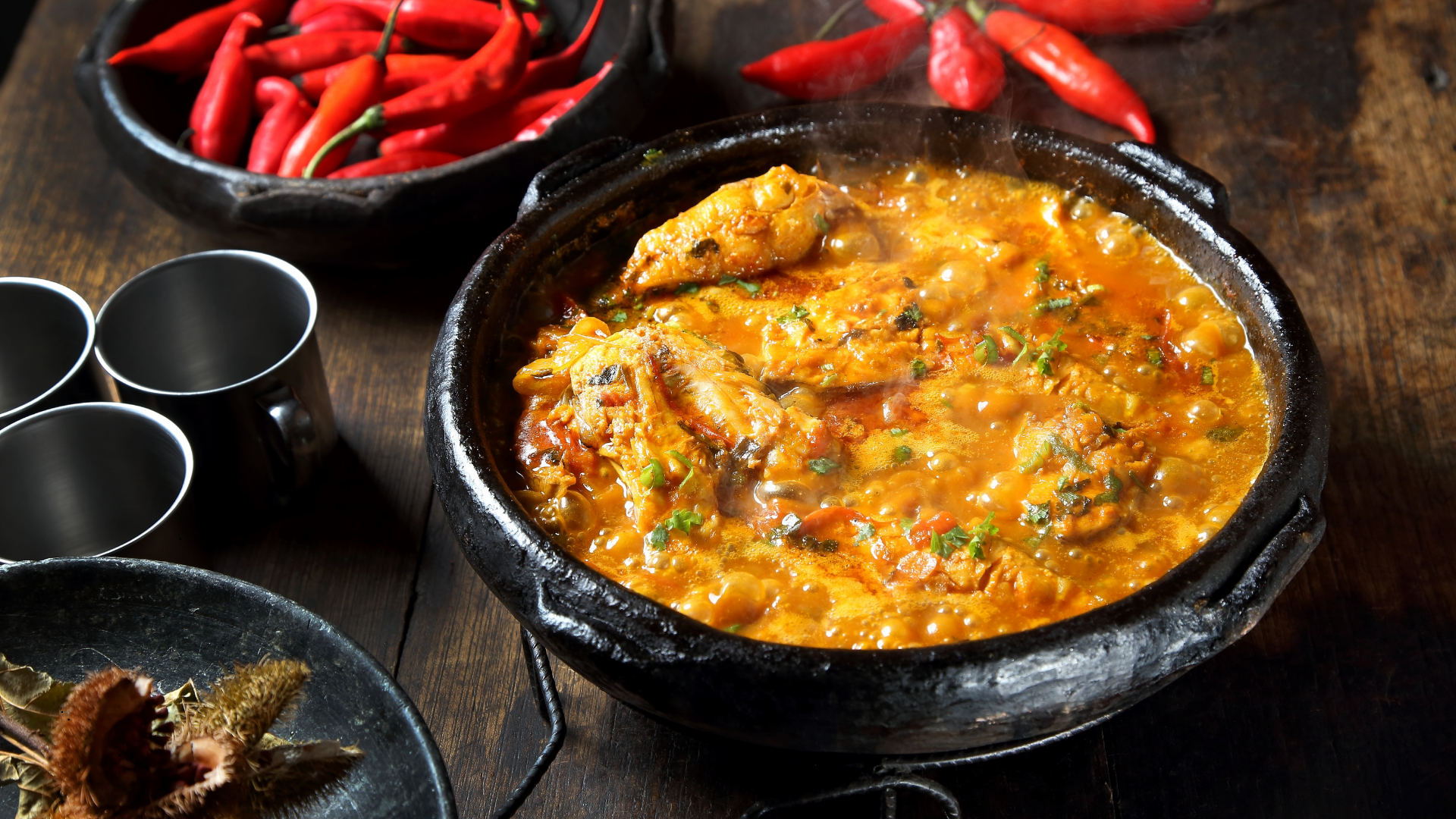 A traditional Mexican clay pot filled with spicy chicken soup garnished with chopped cilantro, with red chili peppers in a bowl and a plate of dried seafood on a wooden table.