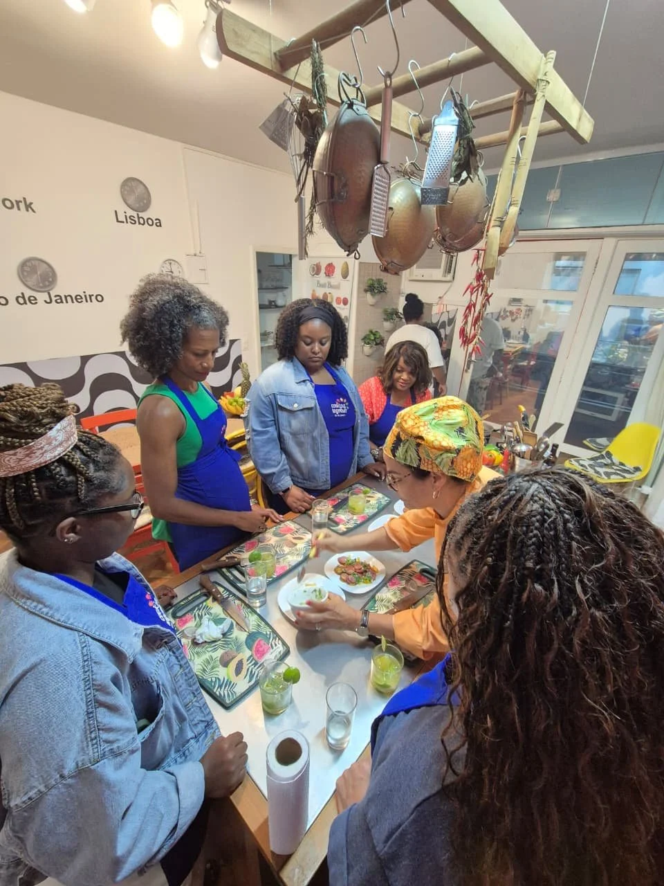 A group of women participating in a cooking class, gathered around a table with food and drinks, in a kitchen with hanging pots and pans.