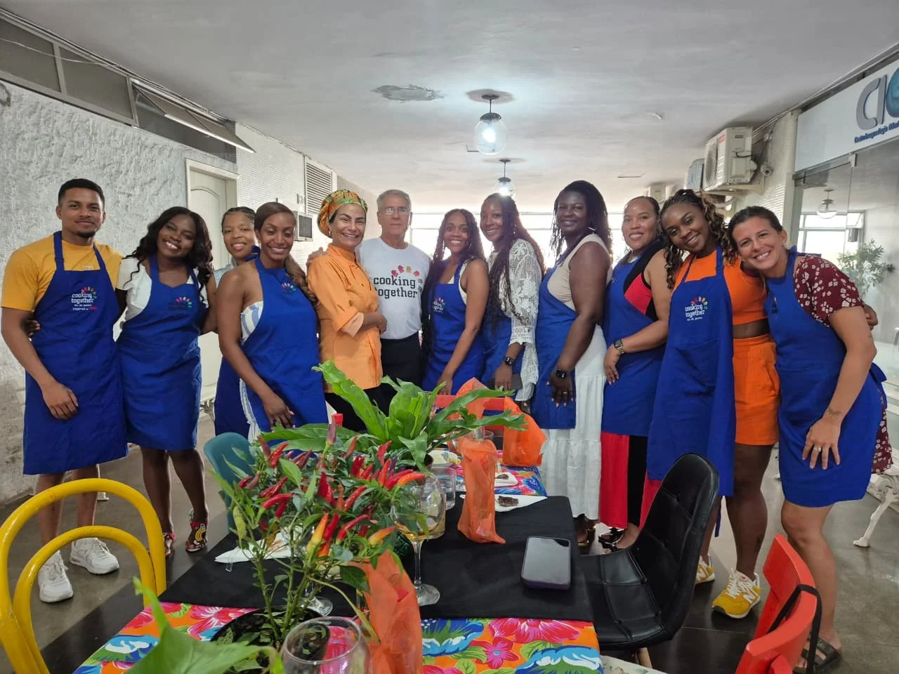 Group of people smiling in a kitchen or cooking class setting, wearing blue aprons, with a table decorated with plants and colorful tablecloths in the foreground.