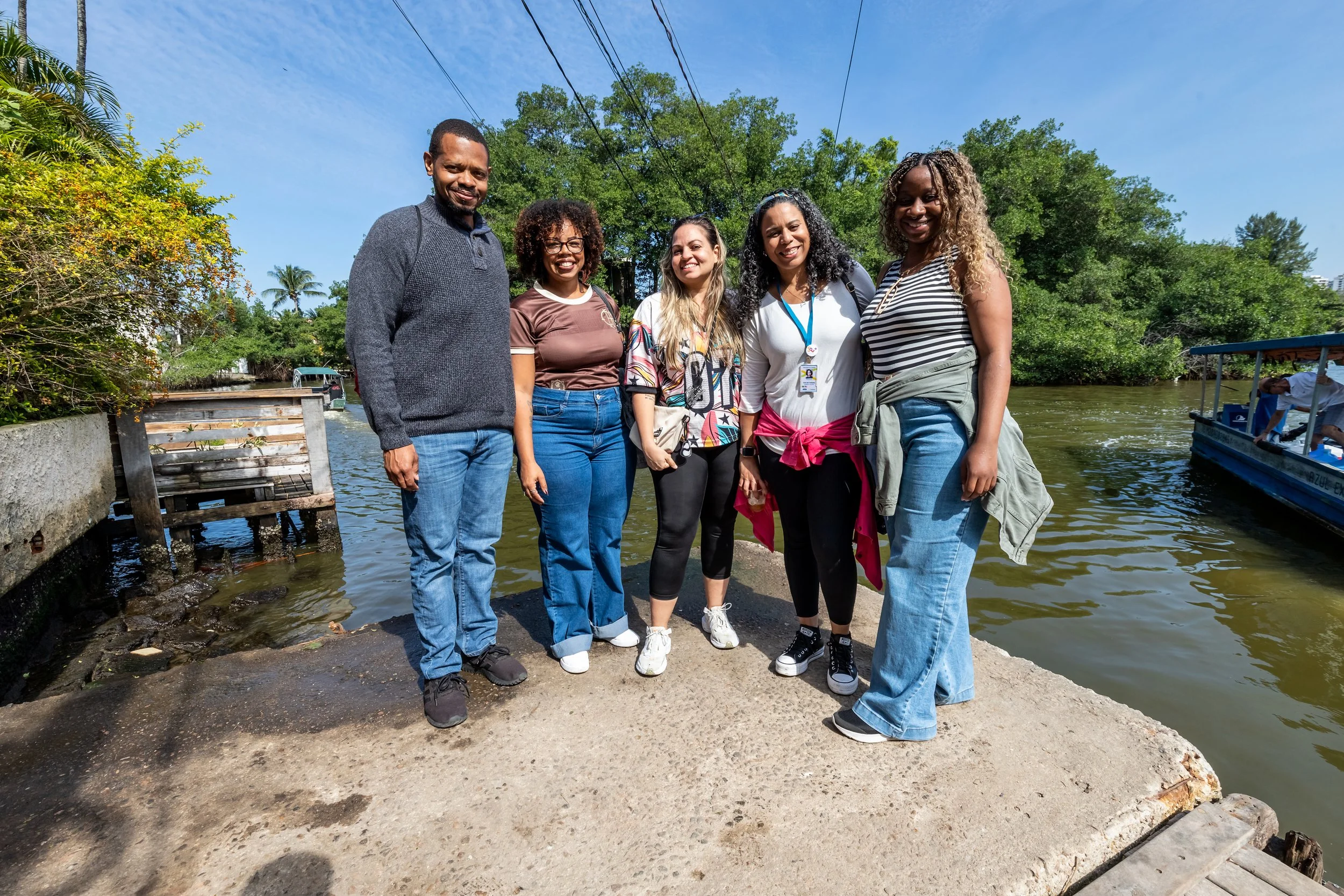 Group of five diverse women and one man standing together outdoors near a river with trees and a boat in the background, smiling at the camera on a sunny day.