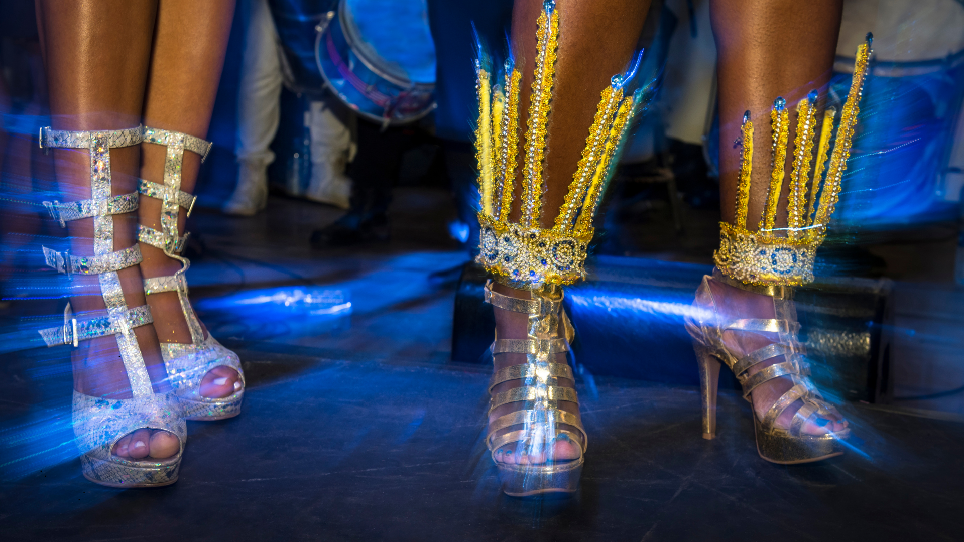 Close-up of a person's legs wearing decorated high-heeled shoes and elaborate, bejeweled leg accessories in gold and silver. The background shows motion blur with blue streaks of light.