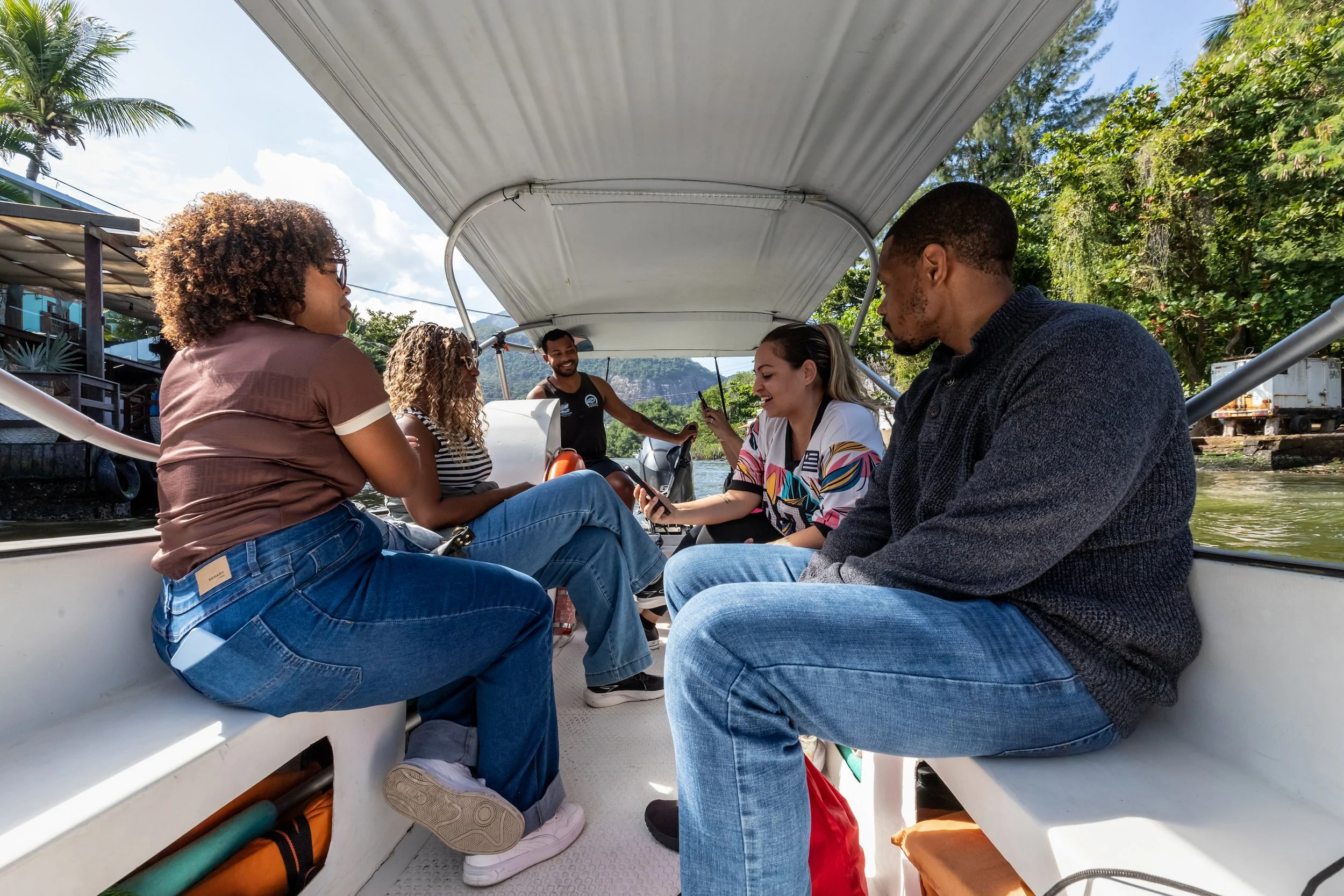 A group of five diverse people sitting in a boat, smiling and socializing during a sunny day with green trees and buildings in the background.