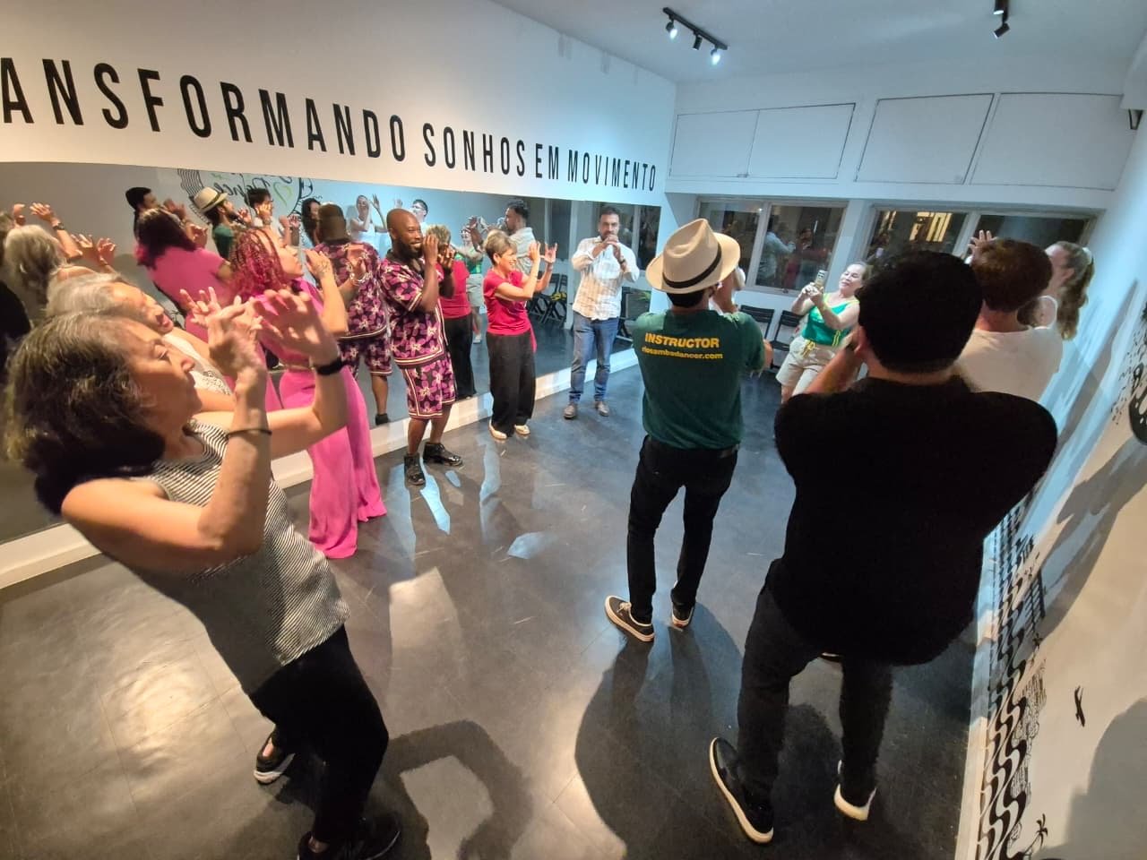 People participating in a dance class or workshop, with some individuals recording or taking photos, in a bright room with large windows and a banner displaying the words 'Transformando Sonhos Em Movimento'.