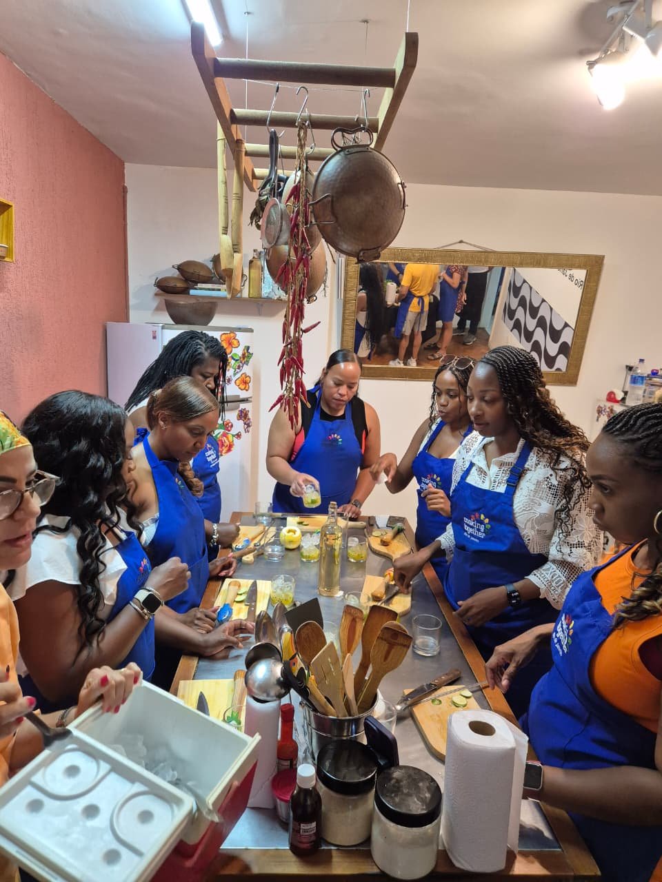 A group of women participating in a cooking class in a kitchen, wearing blue aprons, preparing food at a wooden table with various ingredients and utensils, hanging pots and pans above, and a mirror reflecting other people in the background.