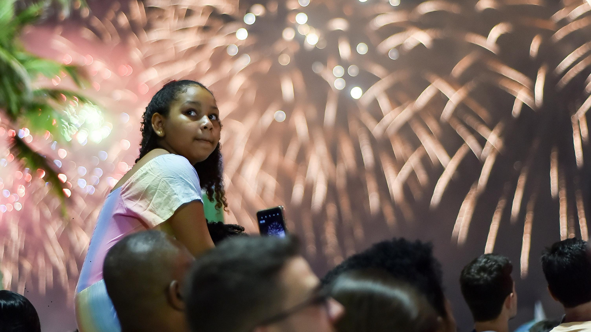 A young girl with braids and earrings sitting among a crowd watching fireworks in the night sky, with colorful bursts of fireworks illuminating the background.