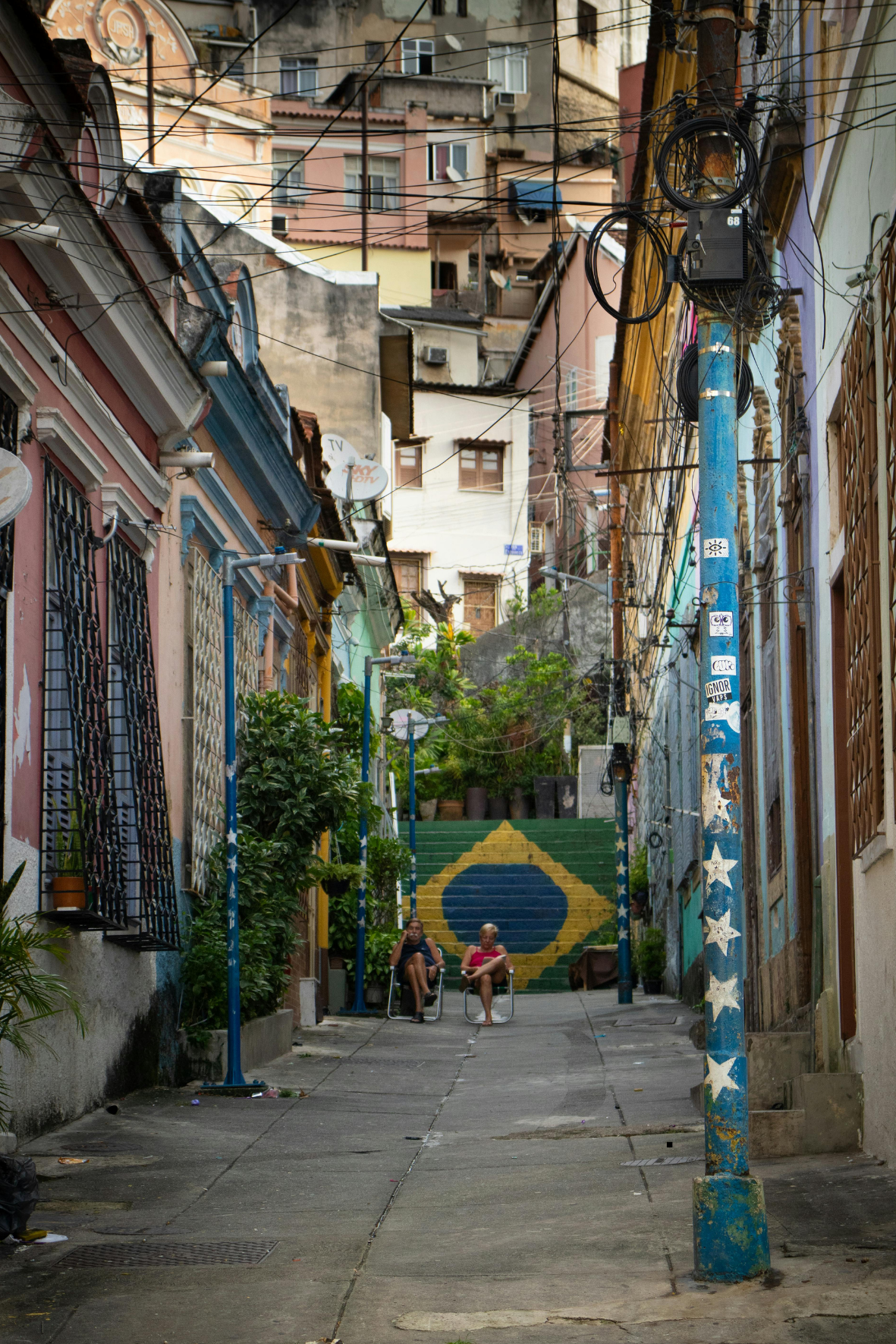 Street scene in a neighborhood with colorful buildings on a hill, electrical wires overhead, a blue pole with star and stripe stickers, stairs painted with the Brazilian flag, and two people sitting on chairs in the foreground.