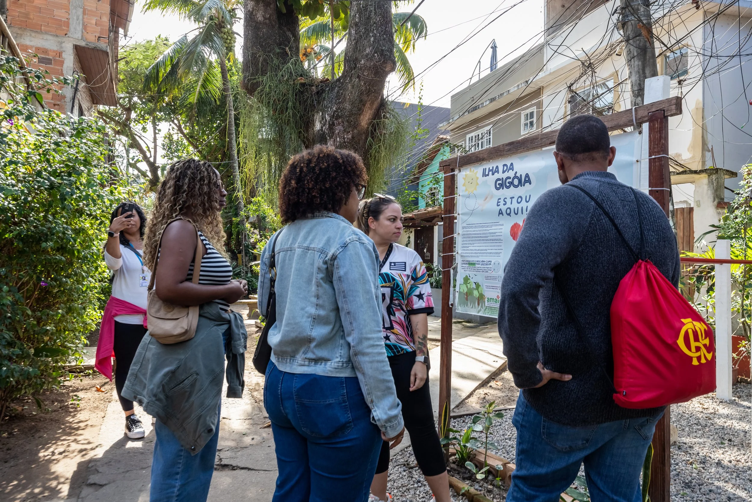 Group of people gathered outdoors in front of a signboard, surrounded by trees and buildings.