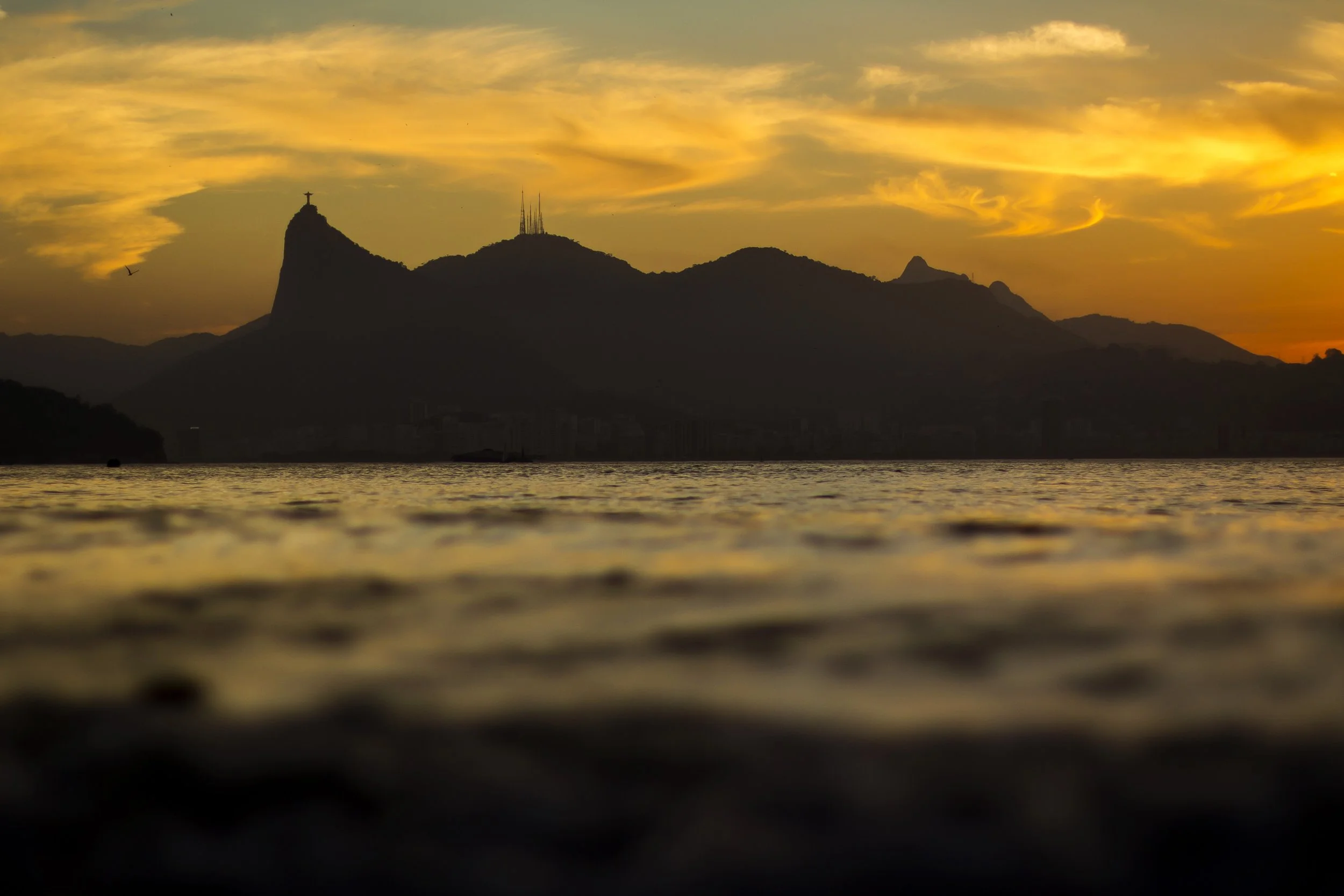 Sunset over a mountainous landscape with the Christ the Redeemer statue on a peak, and communication towers on another, with water in the foreground.