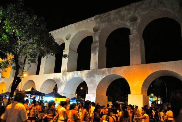 Crowd gathered at night under a large, illuminated historic aqueduct with multiple arches and trees nearby.