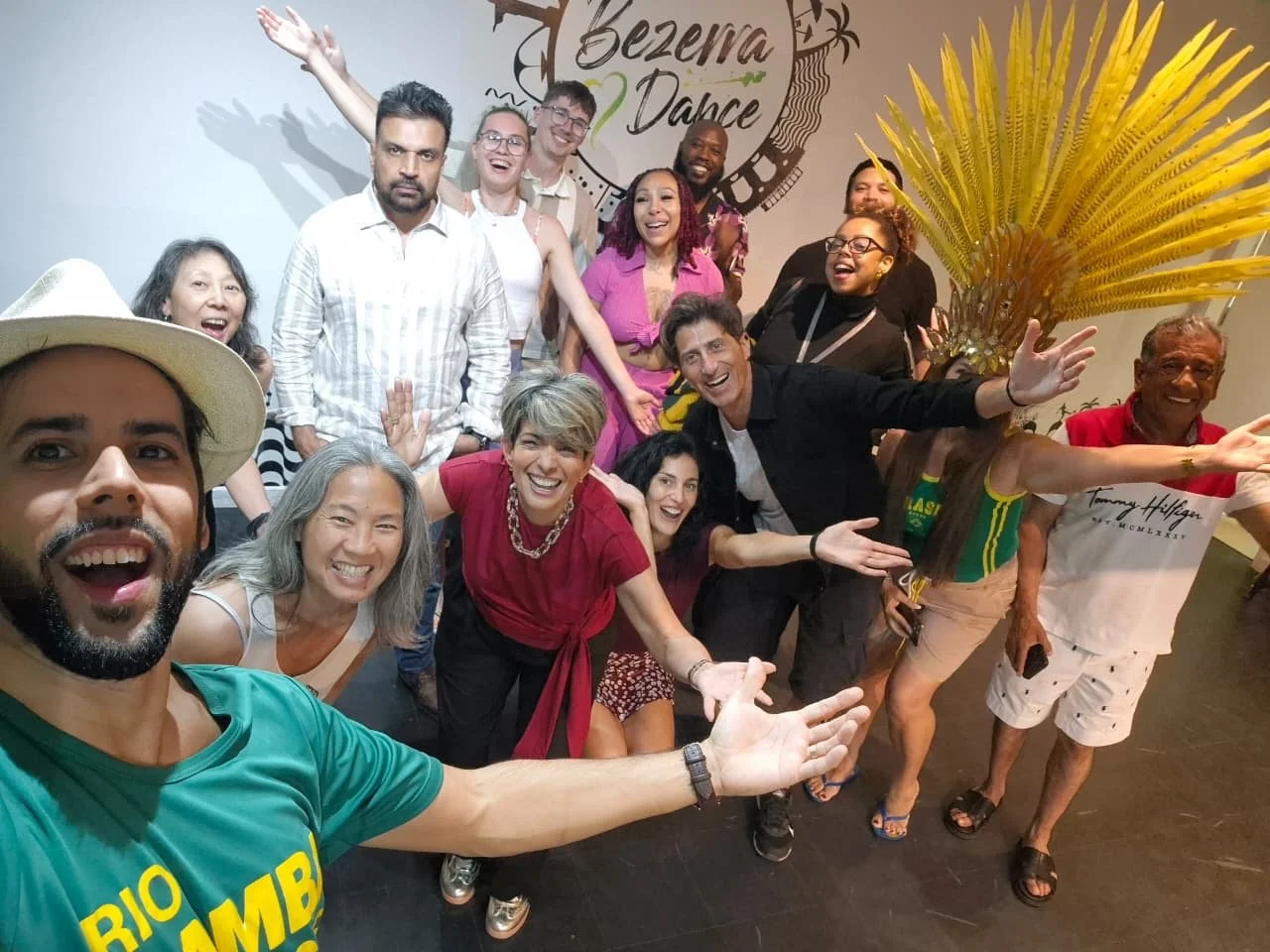 A group of diverse people gathered indoors, smiling and celebrating in front of a sign that reads "Bezerra Dance", with some making playful gestures, and one person wearing a yellow and green shirt from Brazil.