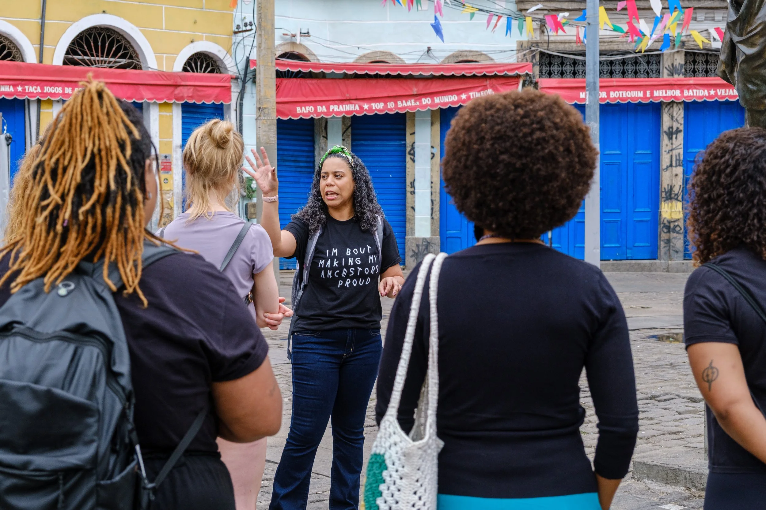 A woman with curly hair wearing a black t-shirt speaking to a group of diverse people in an outdoor setting with colorful flags and closed shops in the background.