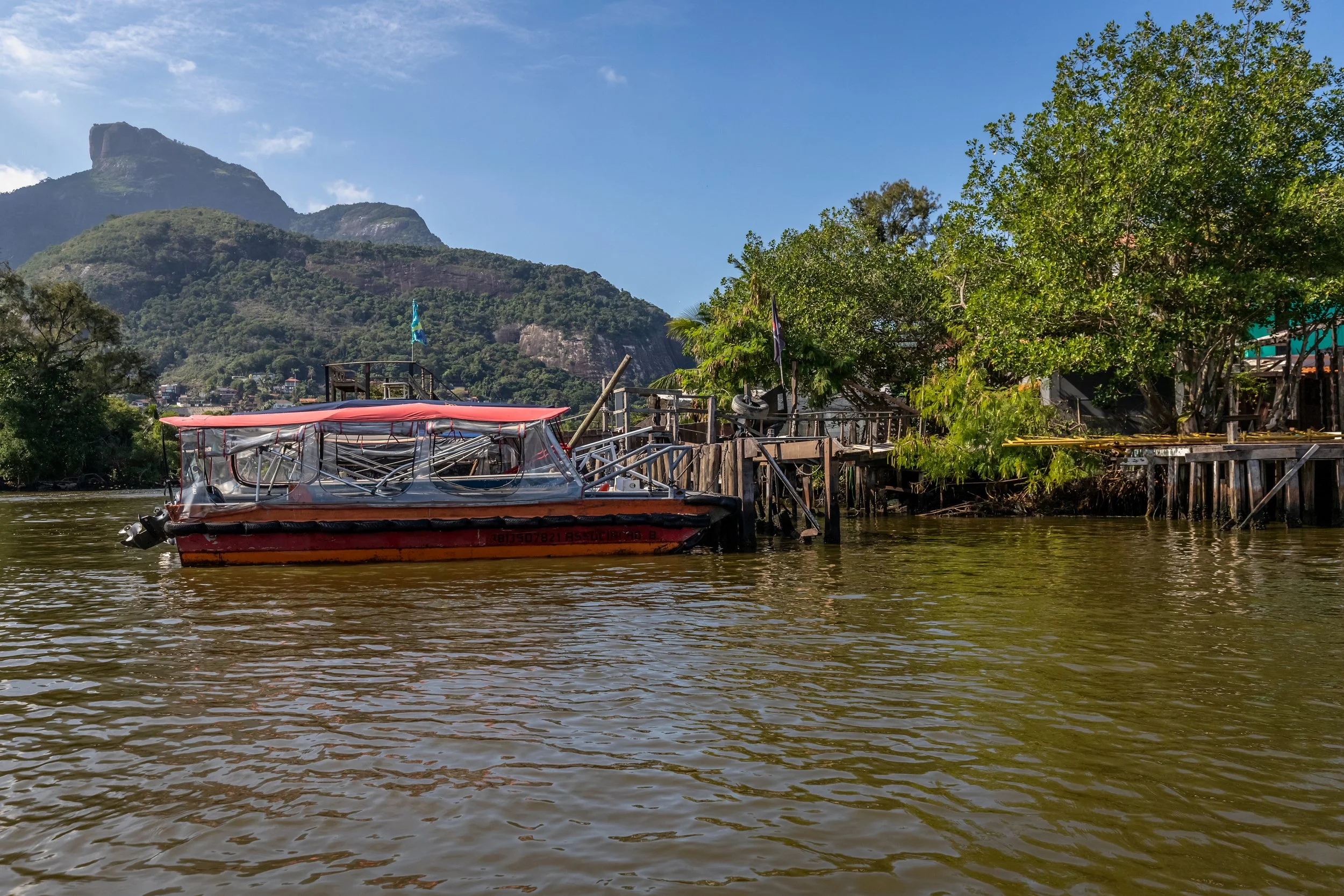 A boat docked along a river with a mountain in the background, green trees on the riverbank, and a wooden dock in a tropical setting.