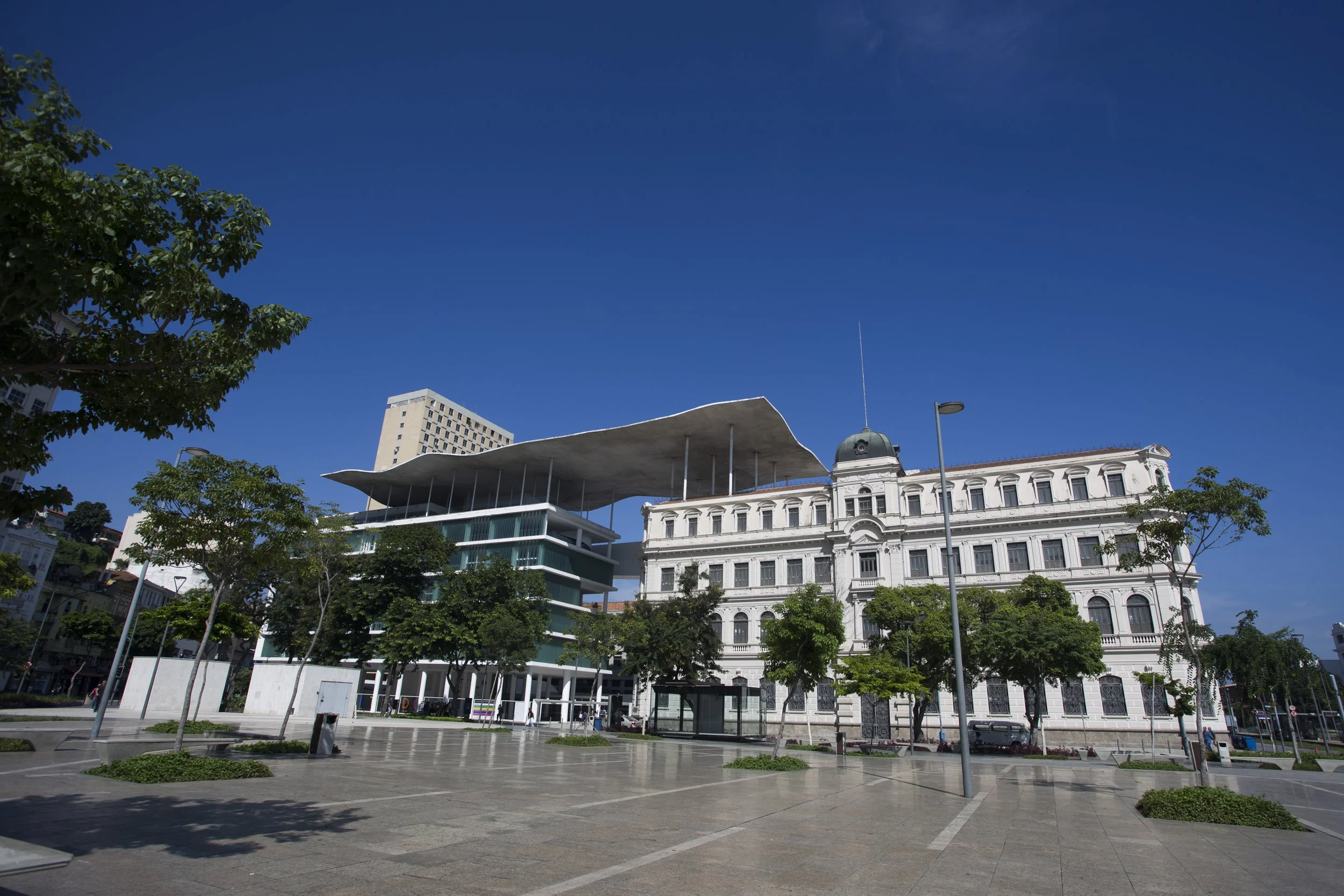 Urban scene with historic white building, modern glass building, green trees, and a blue sky with some clouds.