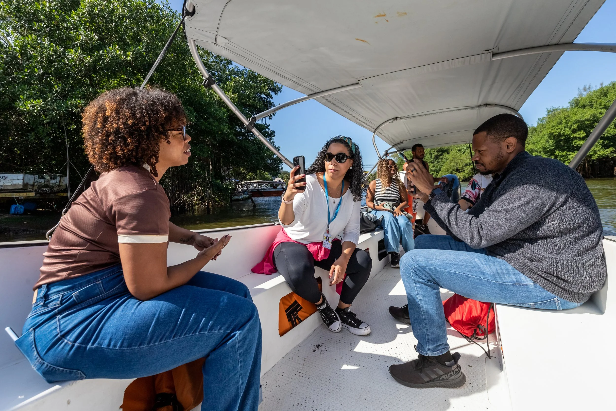 Group of five diverse people sitting on a boat, some looking at their phones, surrounded by green trees and water