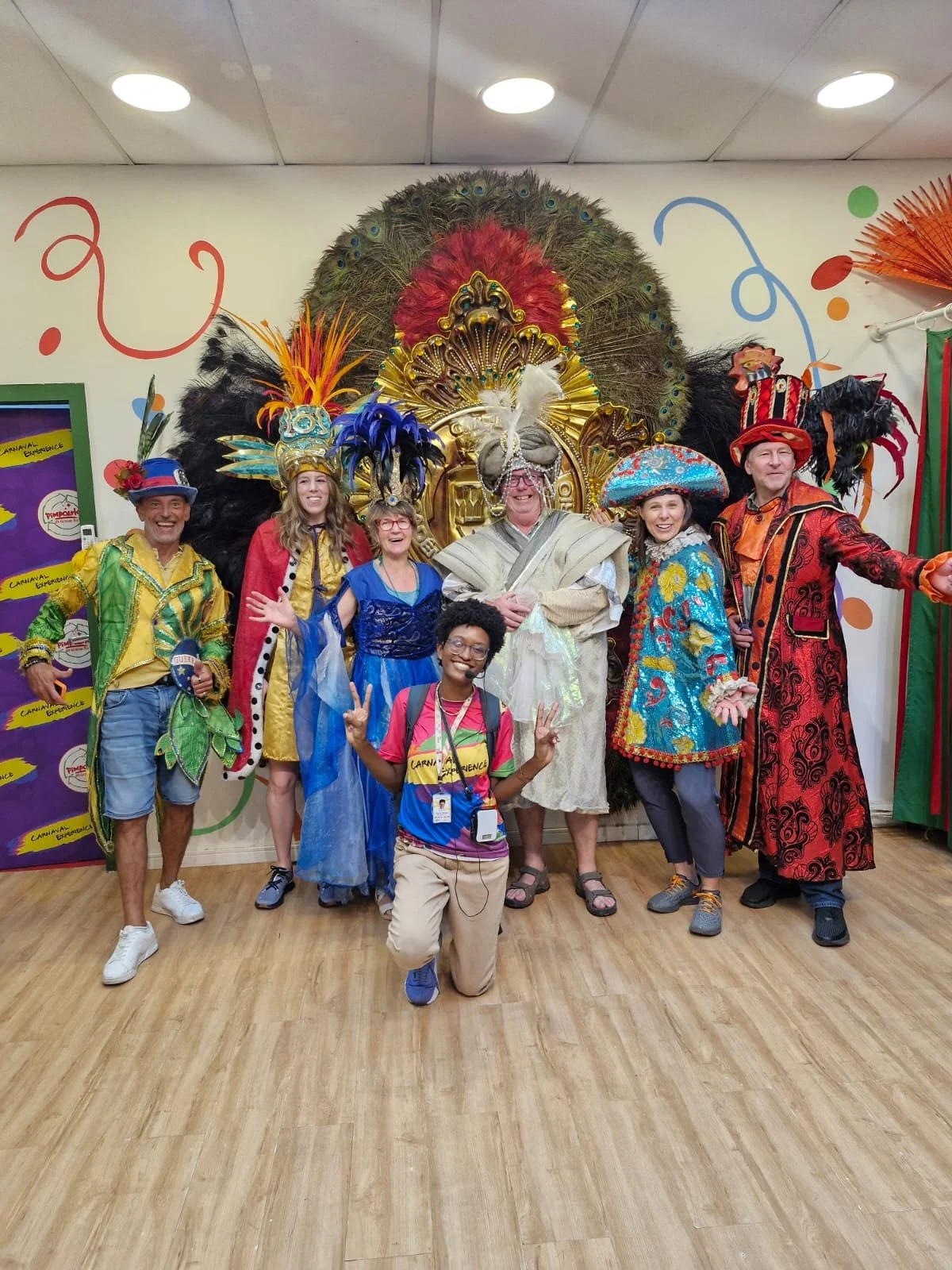 People dressed in colorful carnival costumes posing in front of a decorated backdrop with feathers and decorations, celebrating at a carnival event.