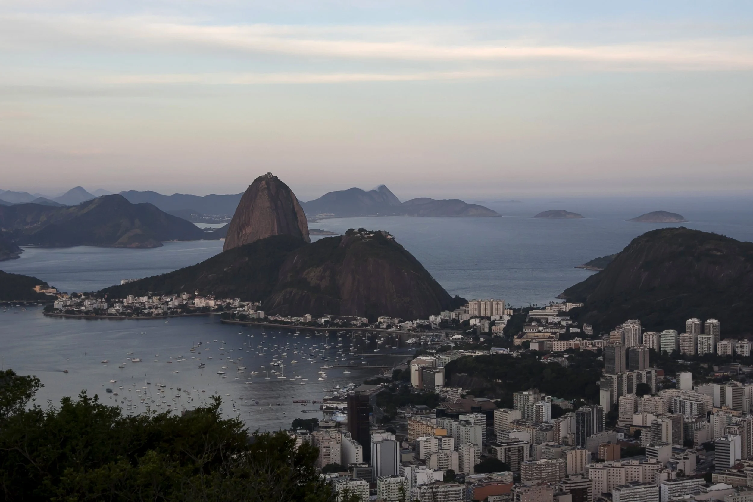 City skyline of Rio de Janeiro with Sugarloaf Mountain, surrounding hills, and bay filled with boats, residential and commercial buildings in the foreground, overcast sky.