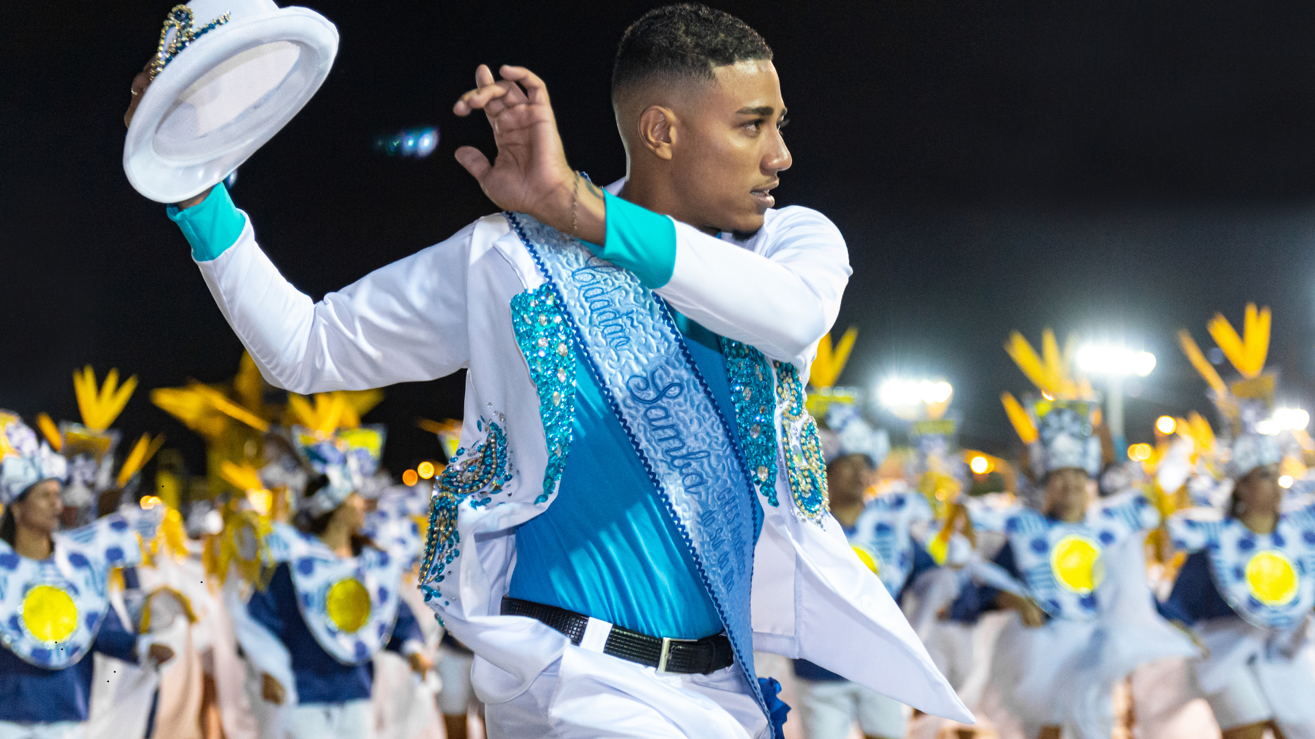 Male performer in a white and blue costume with beads and a sash, waving a white hat during a nighttime parade, with marching band members in blue and yellow uniforms in the background.