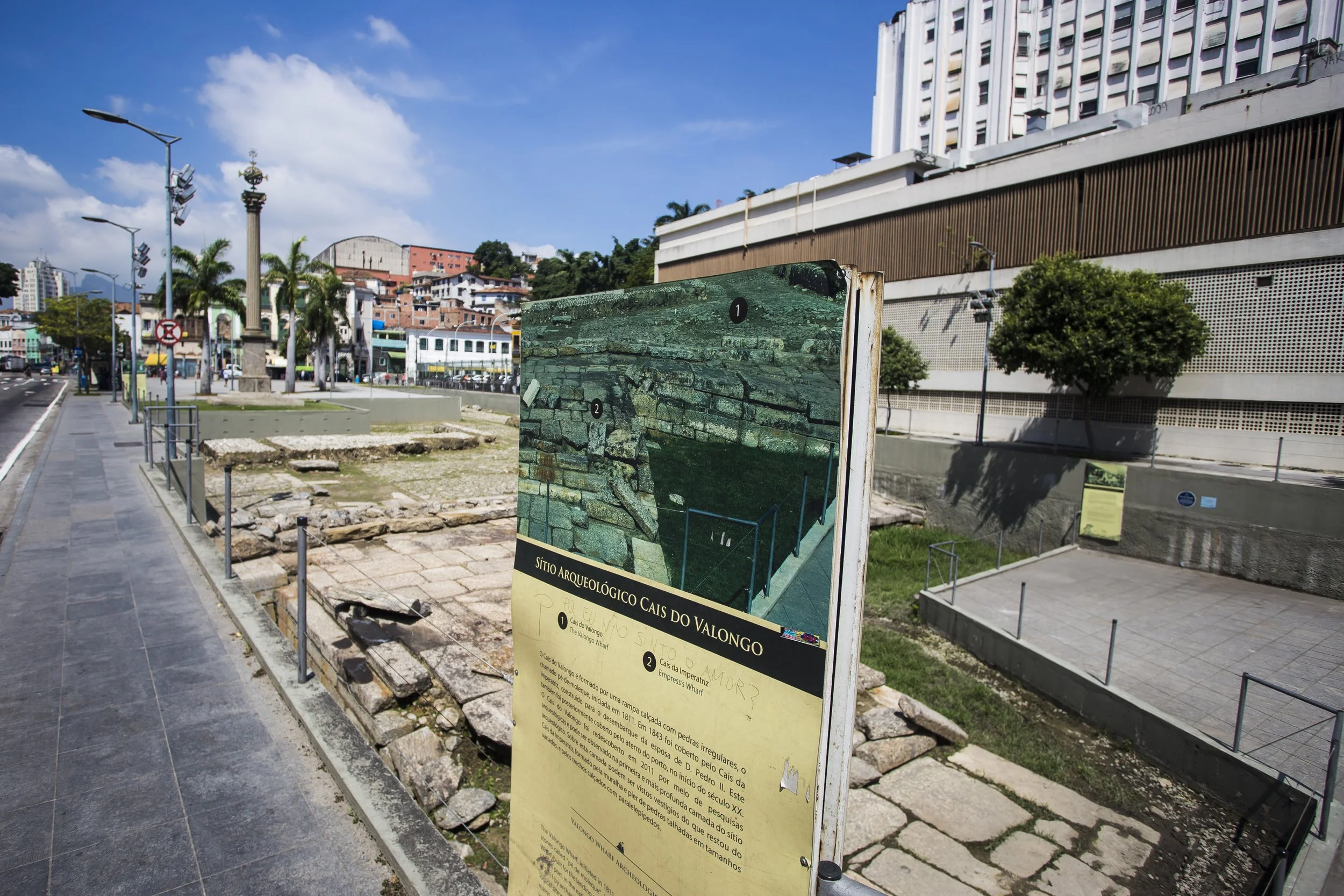 A city street scene with a large archaeological sign in the foreground, palm trees, modern buildings, and a clear blue sky. The archaeological site features ancient stone walls and ruins, with informational panels describing the site.