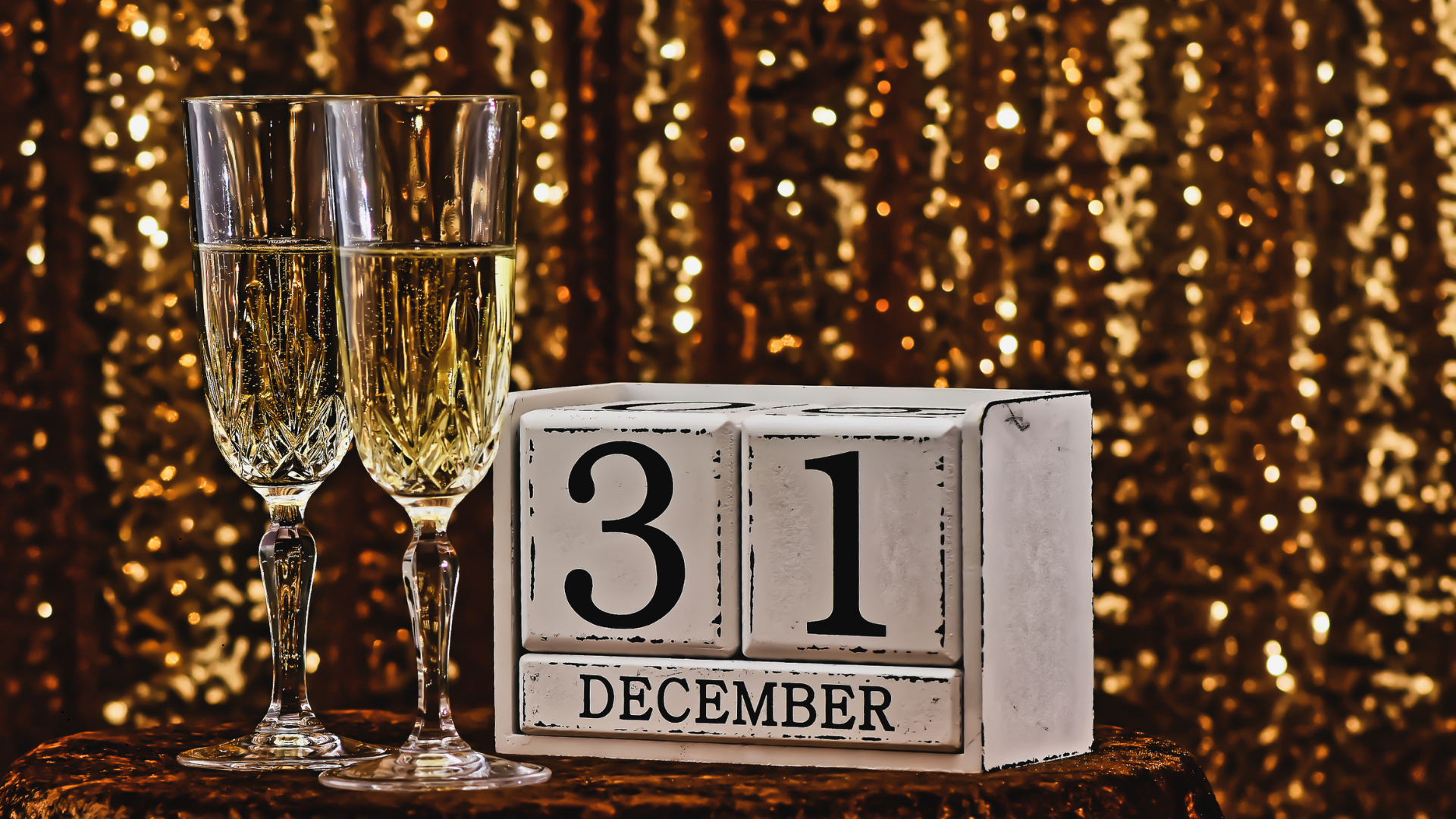 Two champagne flutes filled with champagne next to a white block calendar showing December 31, with a sparkly gold background.