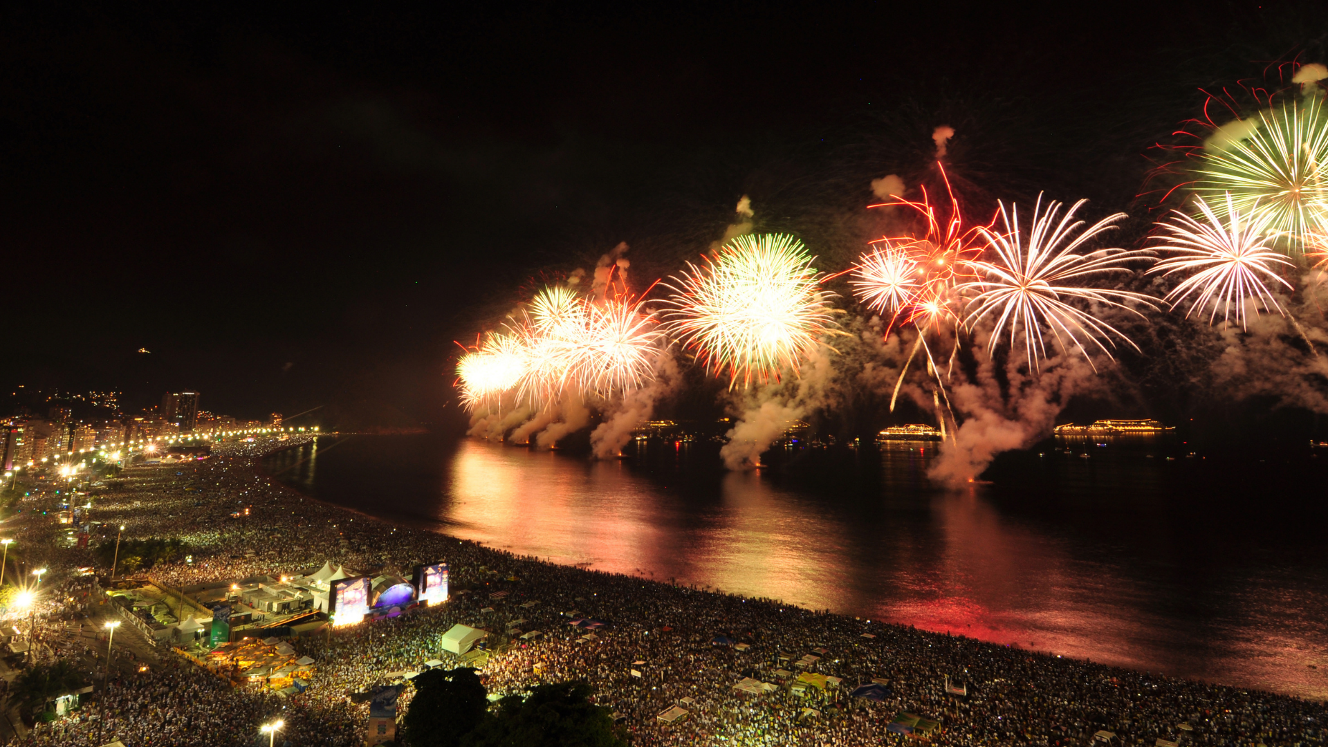 Fireworks display over a beach at night, with a large crowd of people on the shoreline and city lights in the background.