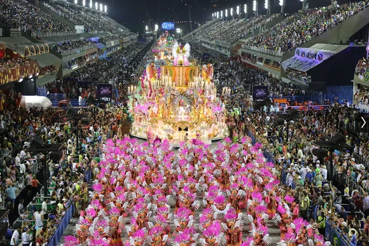 Large parade with a decorated float at a nighttime event, surrounded by thousands of spectators and participants in colorful costumes.