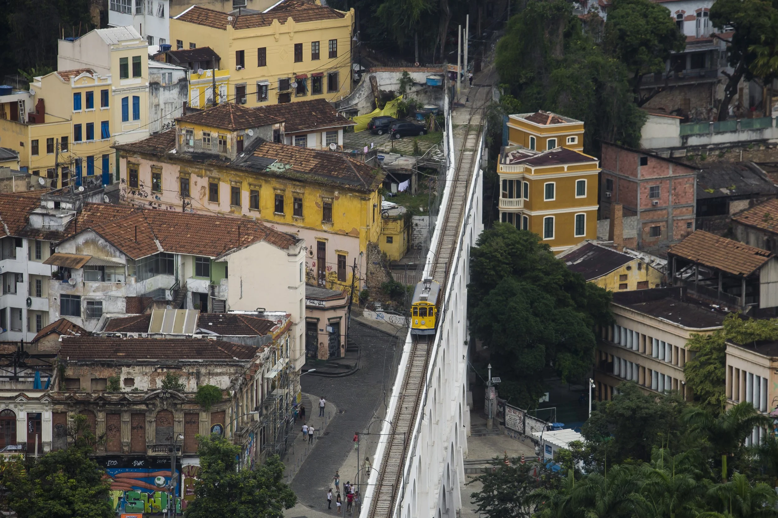 A city scene with a variety of colorful old and new buildings, a yellow streetcar on elevated tracks, and a cobblestone street below with pedestrians walking.