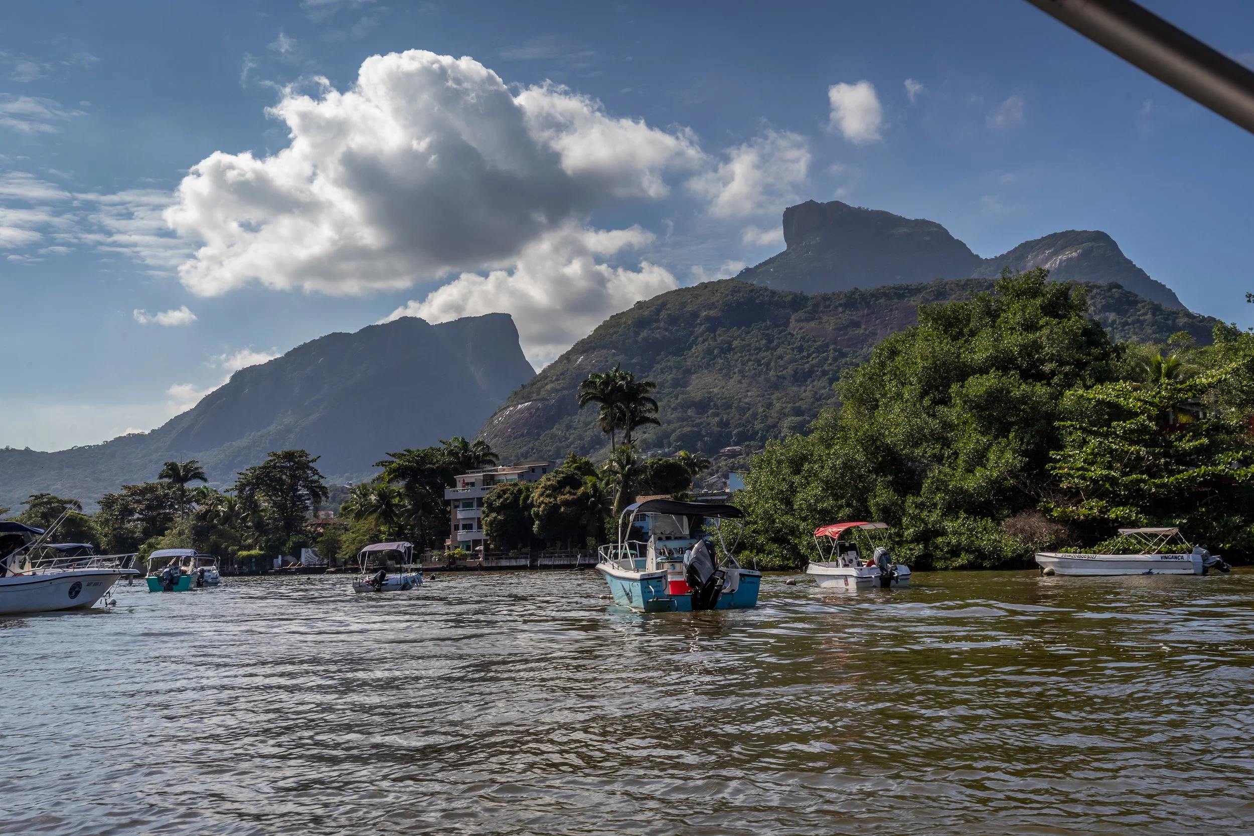 Marina with boats docked on the water, lush green trees, and mountains in the background under a blue sky with scattered clouds.