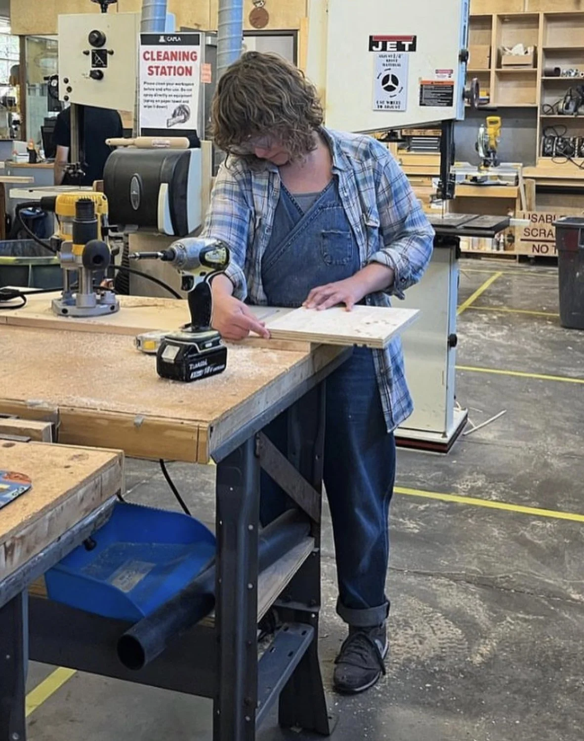Rachel is working in a woodworking shop, measuring a piece of wood on a workbench with tools and machinery around.