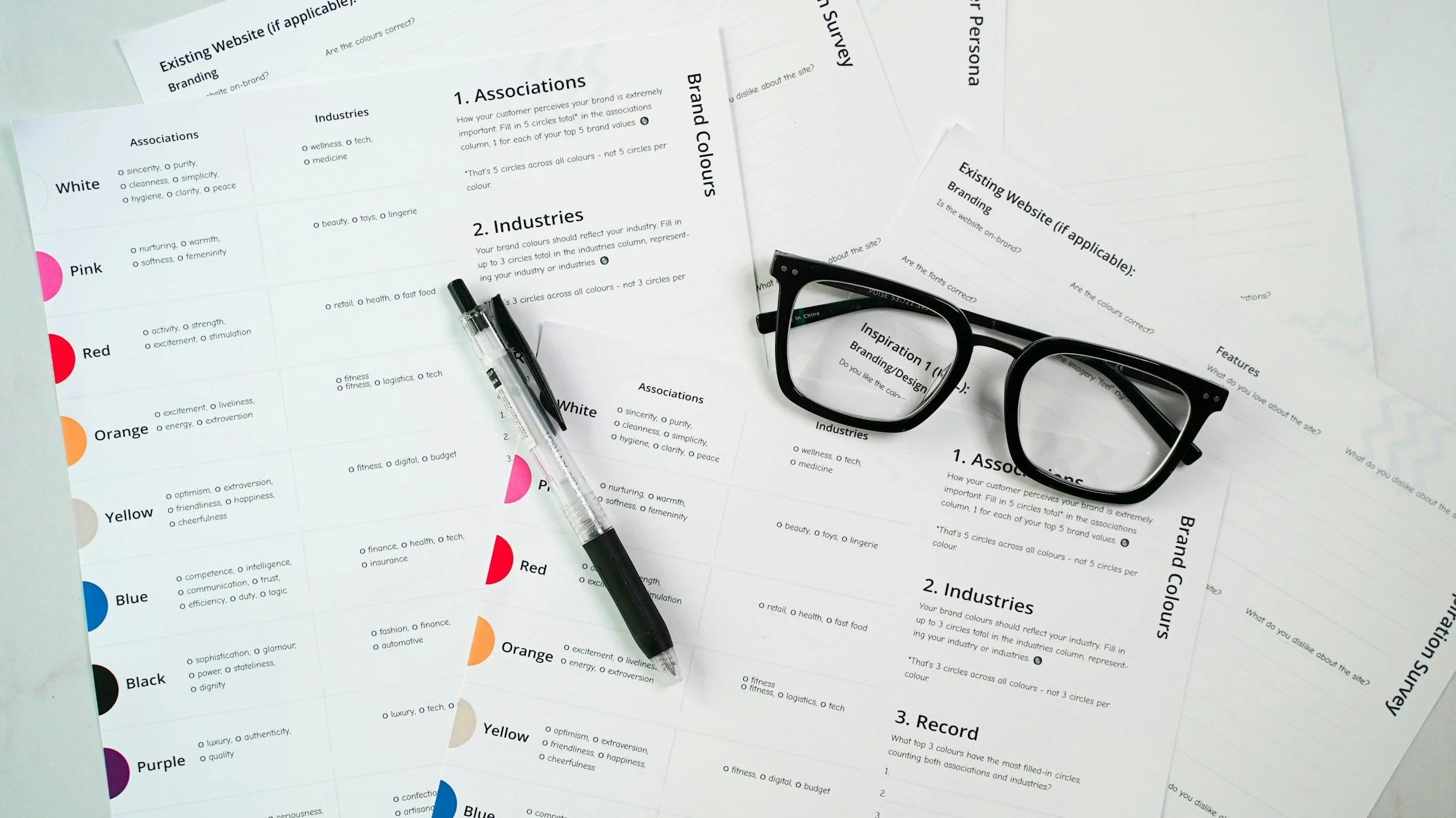 Desk with eyeglasses, black pen, and printed sheets of paper with text about brand colors and associations, labels in different colors, and survey questions.