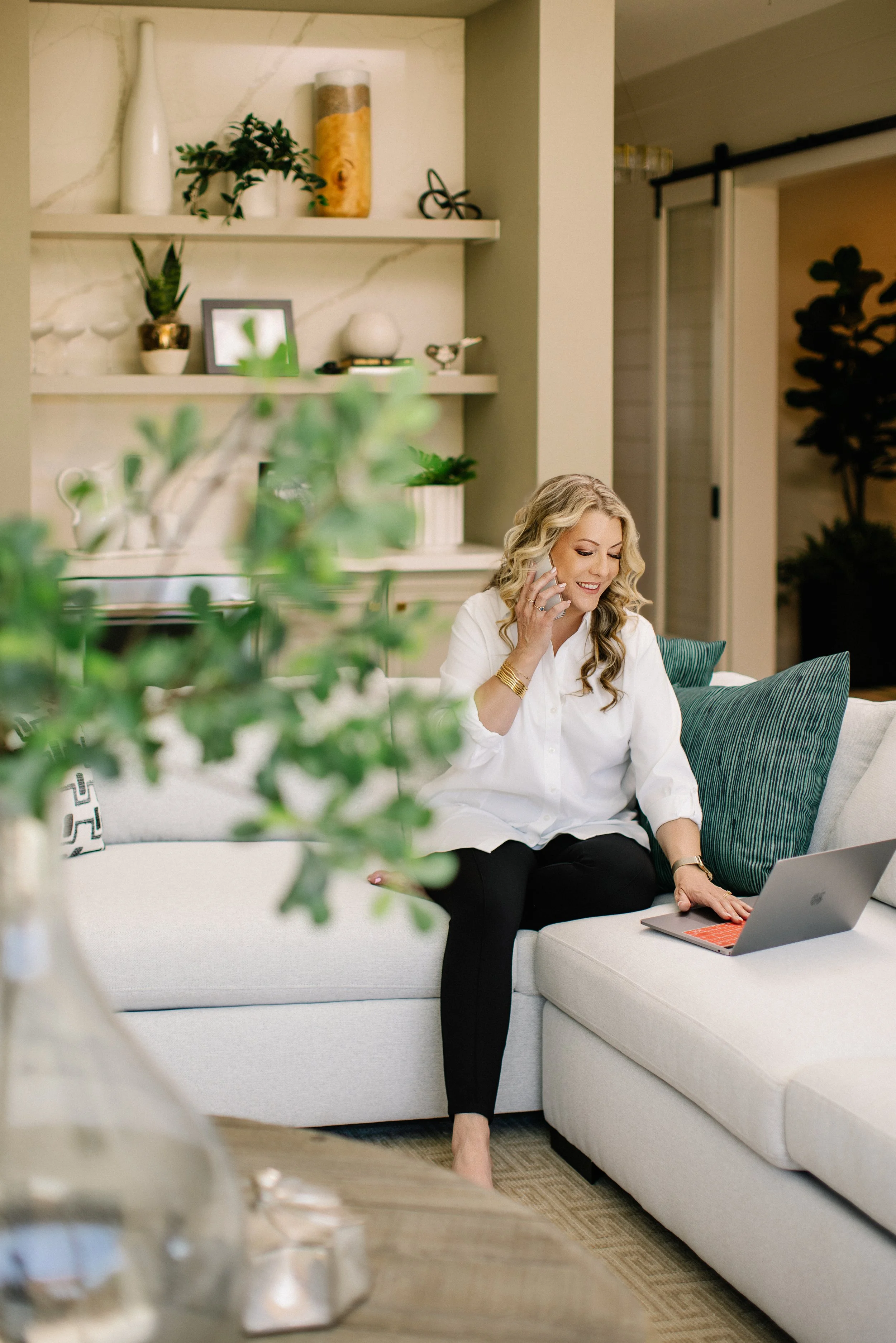 A woman sitting on a cream-colored sofa talking on her cellphone and using a laptop, with a green pillow behind her and a blurred plant in foreground.