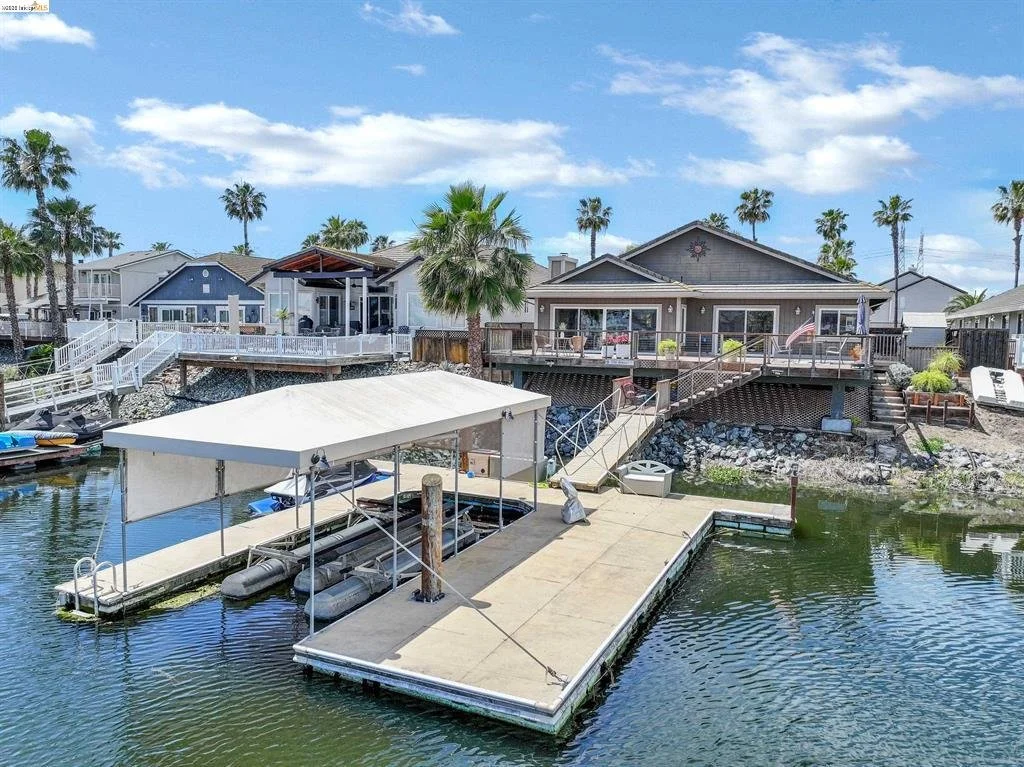 A waterfront scene showing a dock with a covered boat lift holding inflatable boats, and houses with decks and palm trees in the background under a partly cloudy sky.