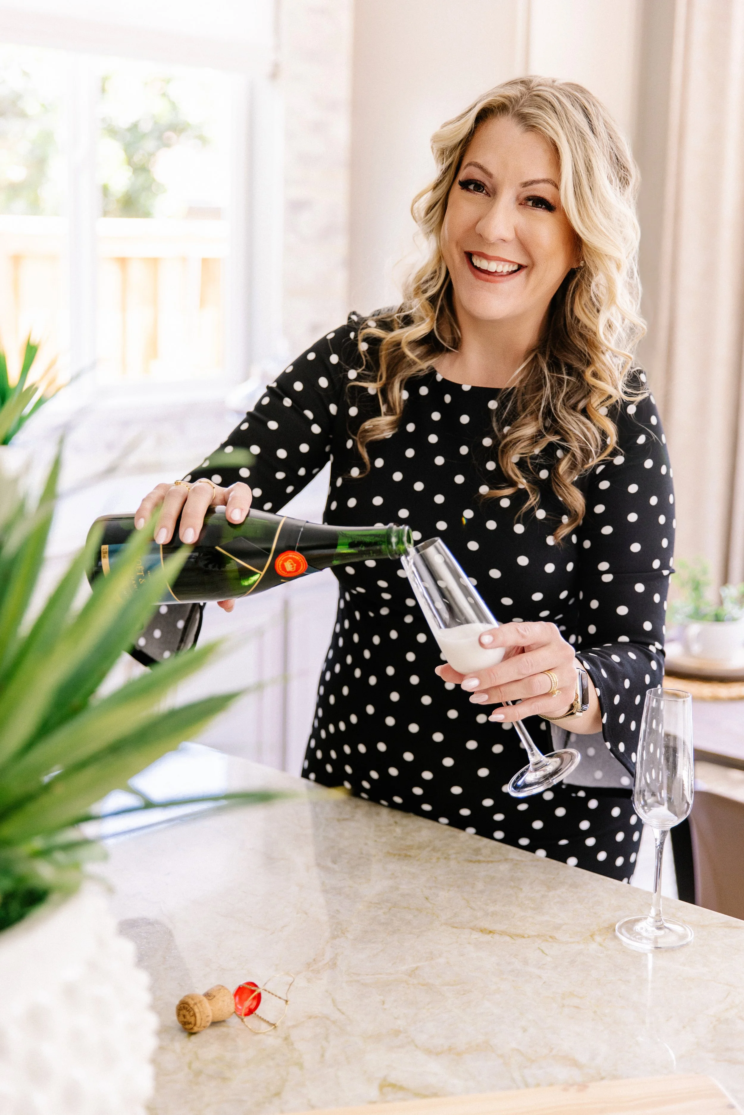 A woman with blonde, curly hair smiling as she pours champagne into a flute at a kitchen counter, wearing a black dress with white polka dots.
