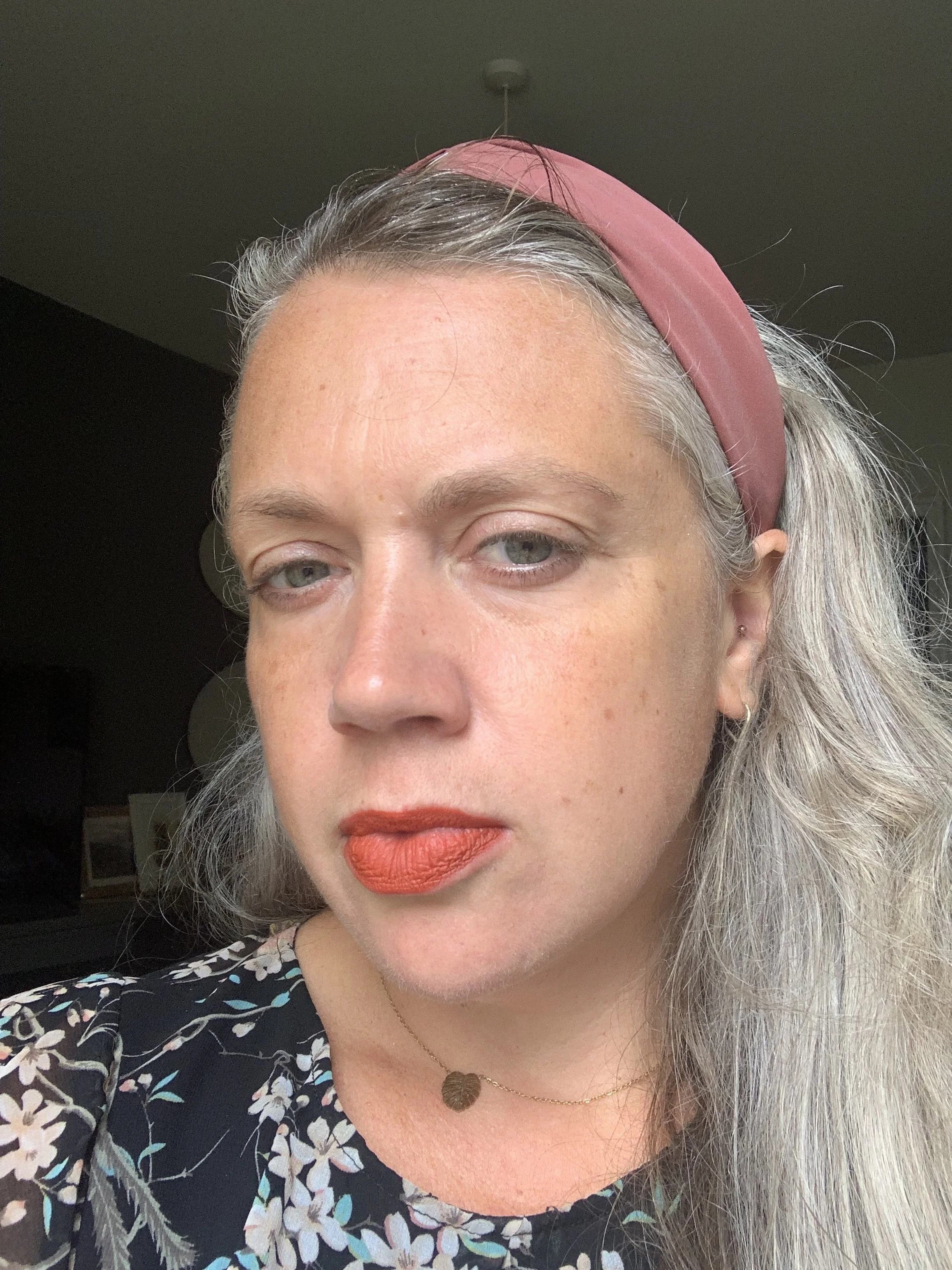 A woman with long gray hair wearing a pink headband, floral black top, and orange lipstick, looking slightly to the side.