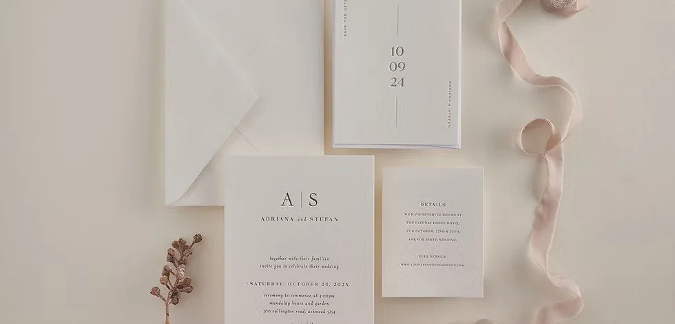 Flat lay of white wedding invitation suite with envelope, date card, details card, and ribbon on a neutral background.