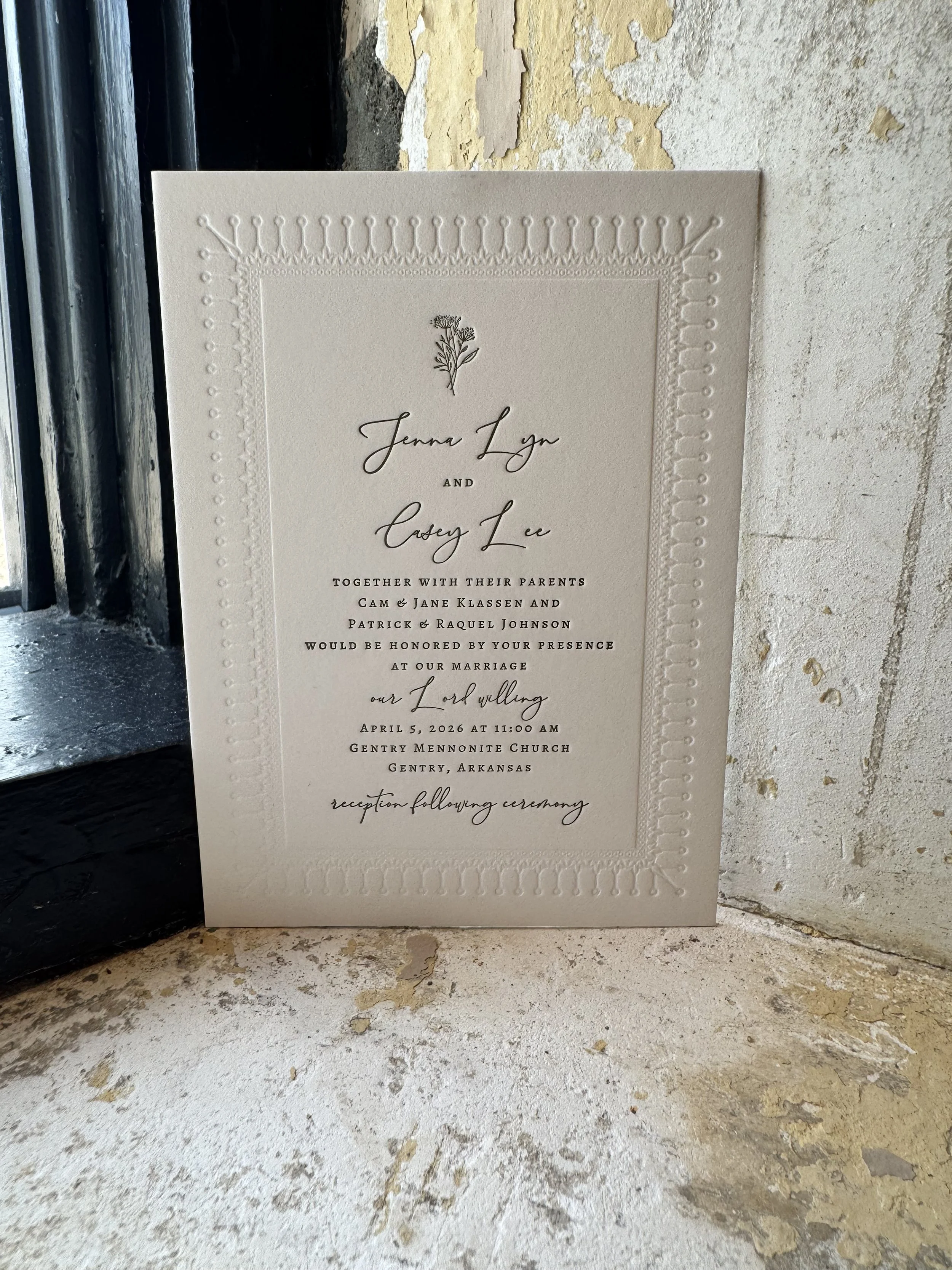 A wedding invitation card with elegant script and floral design, placed against a rustic wall.