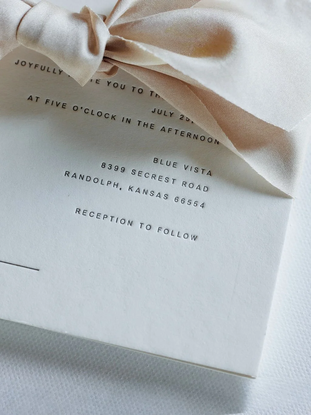 Close-up of a wedding invitation with a pink ribbon and elegant black text, including details of date, time, and location.