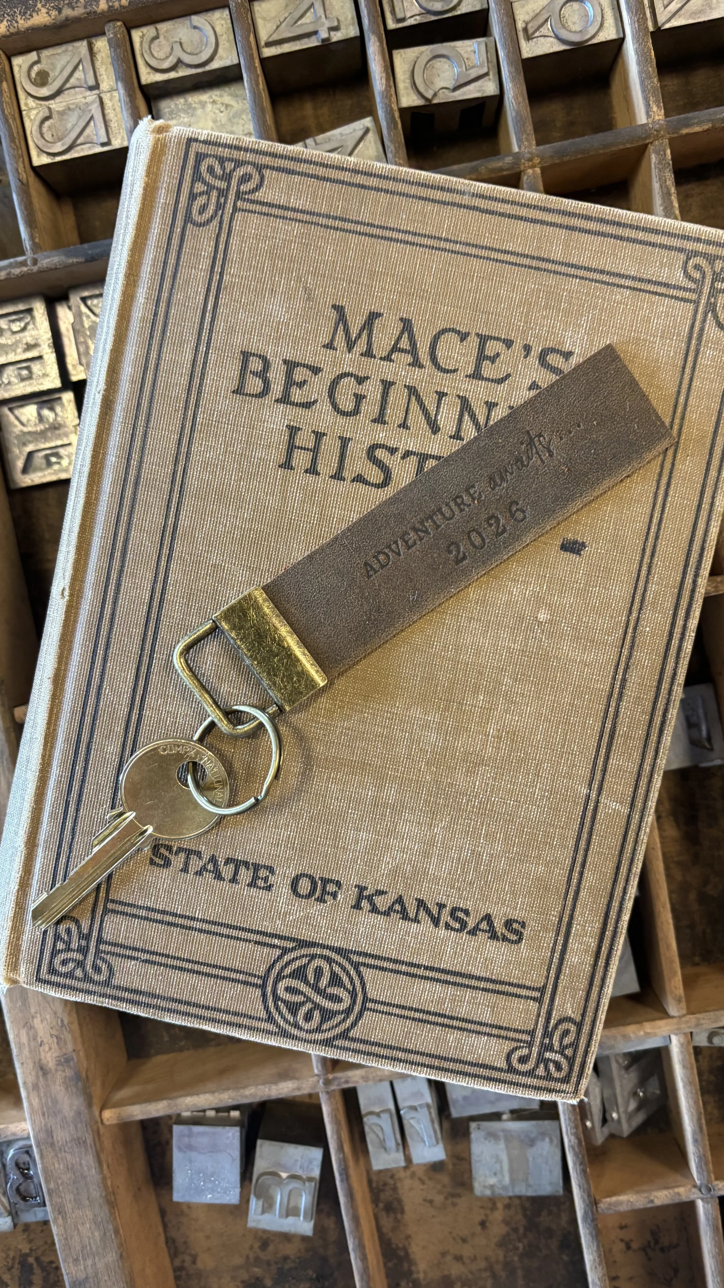A book titled 'Mace's Beginning History' with a sleeve tag labeled 'Adventure & Outdoors 2026'. A keychain with keys is hanging from the sleeve. The background features a wooden box filled with metal letter stamps.