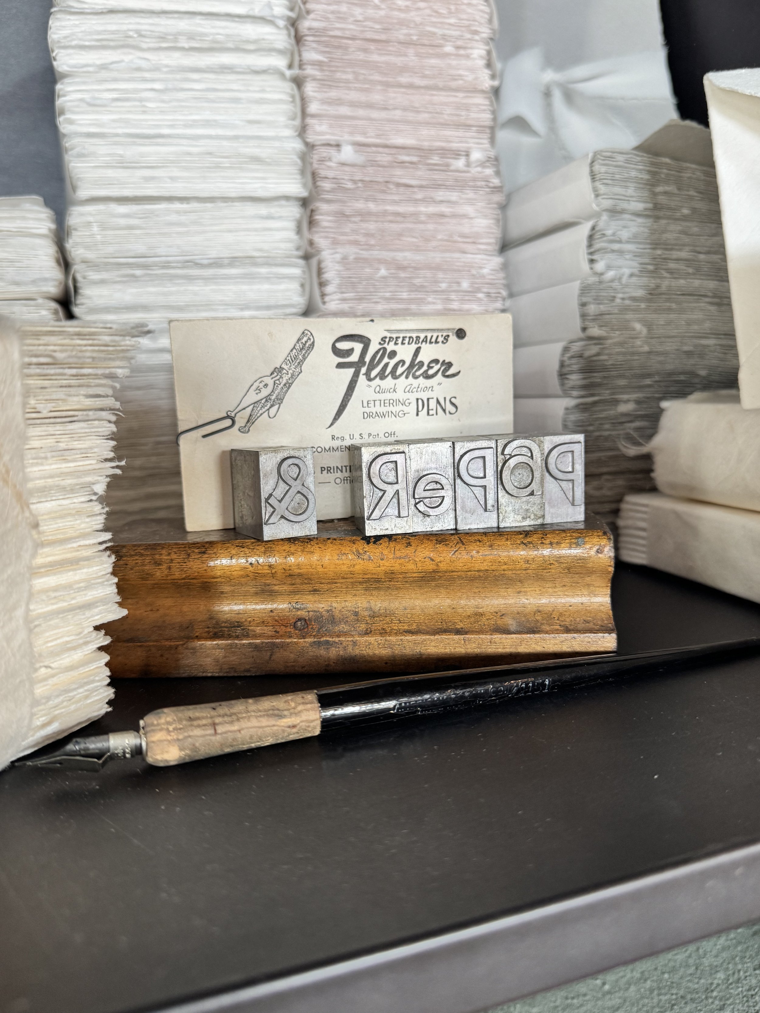 Close-up of rubber stamps, a pen, and a packet of vintage letterpress printing blocks labeled 'Speedball's Flicker' and 'Lettering Drawing Pens' on a wooden surface.