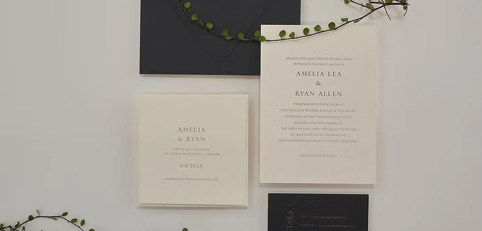 Wedding invitation suite with white and black cards, surrounded by greenery.