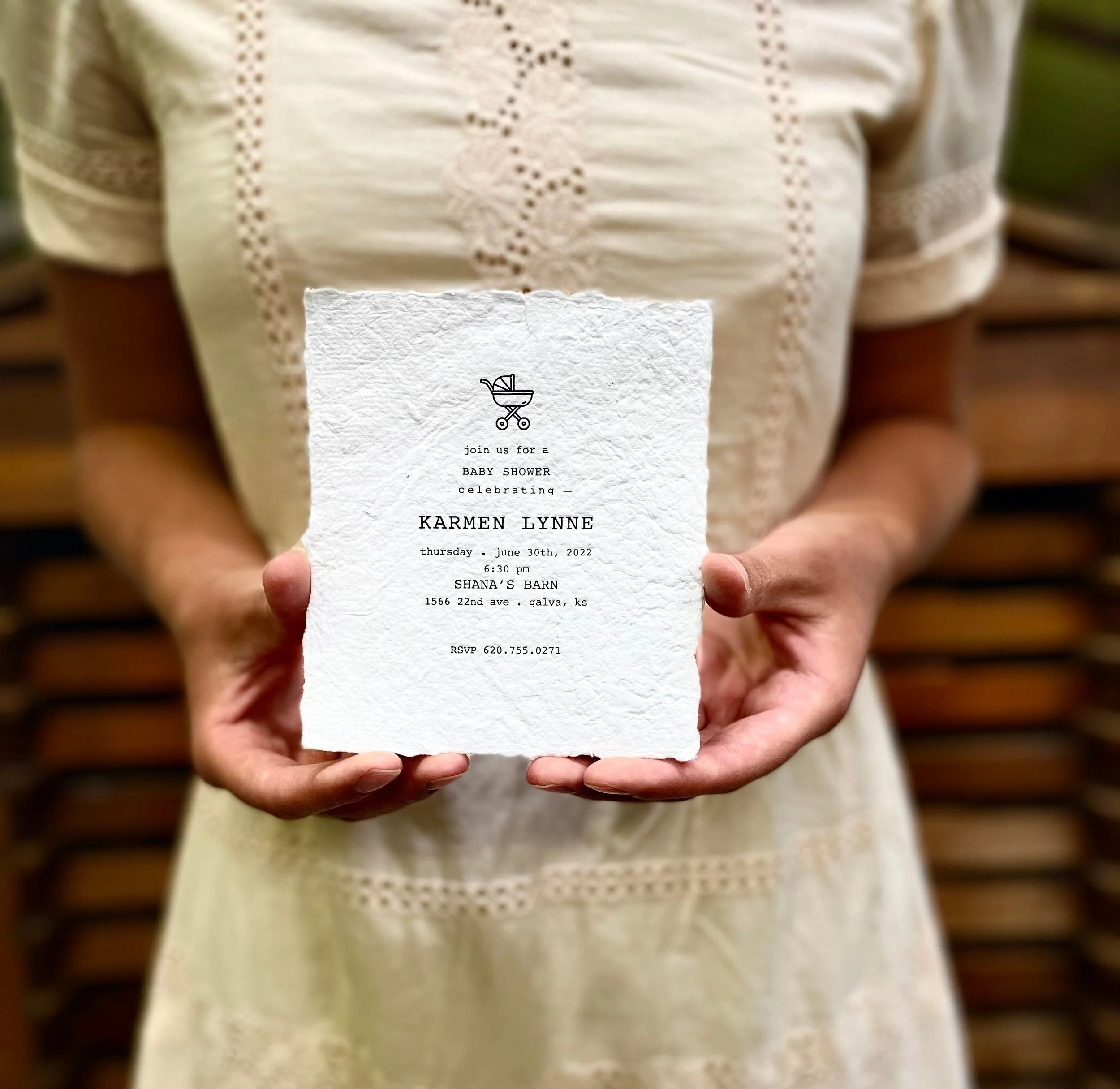 A person holding a white invitation card with black text and a small graphic of a baby stroller, for a baby shower celebration. The invitation is partially visible with details about the event, including the date, time, location, and RSVP information