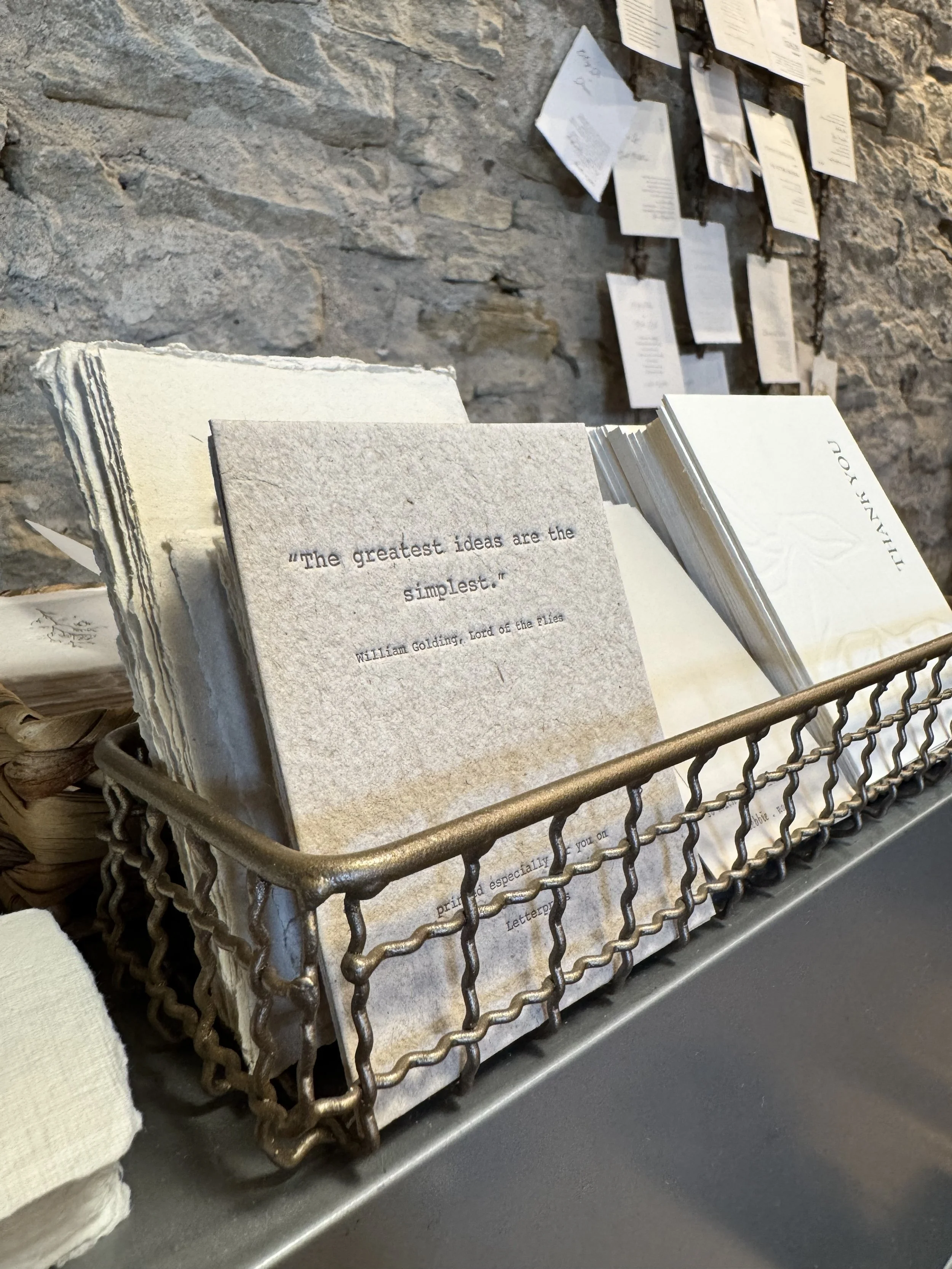 Open wire basket filled with folded paper napkins, placed on a metal counter in front of a stone wall with hanging papers.