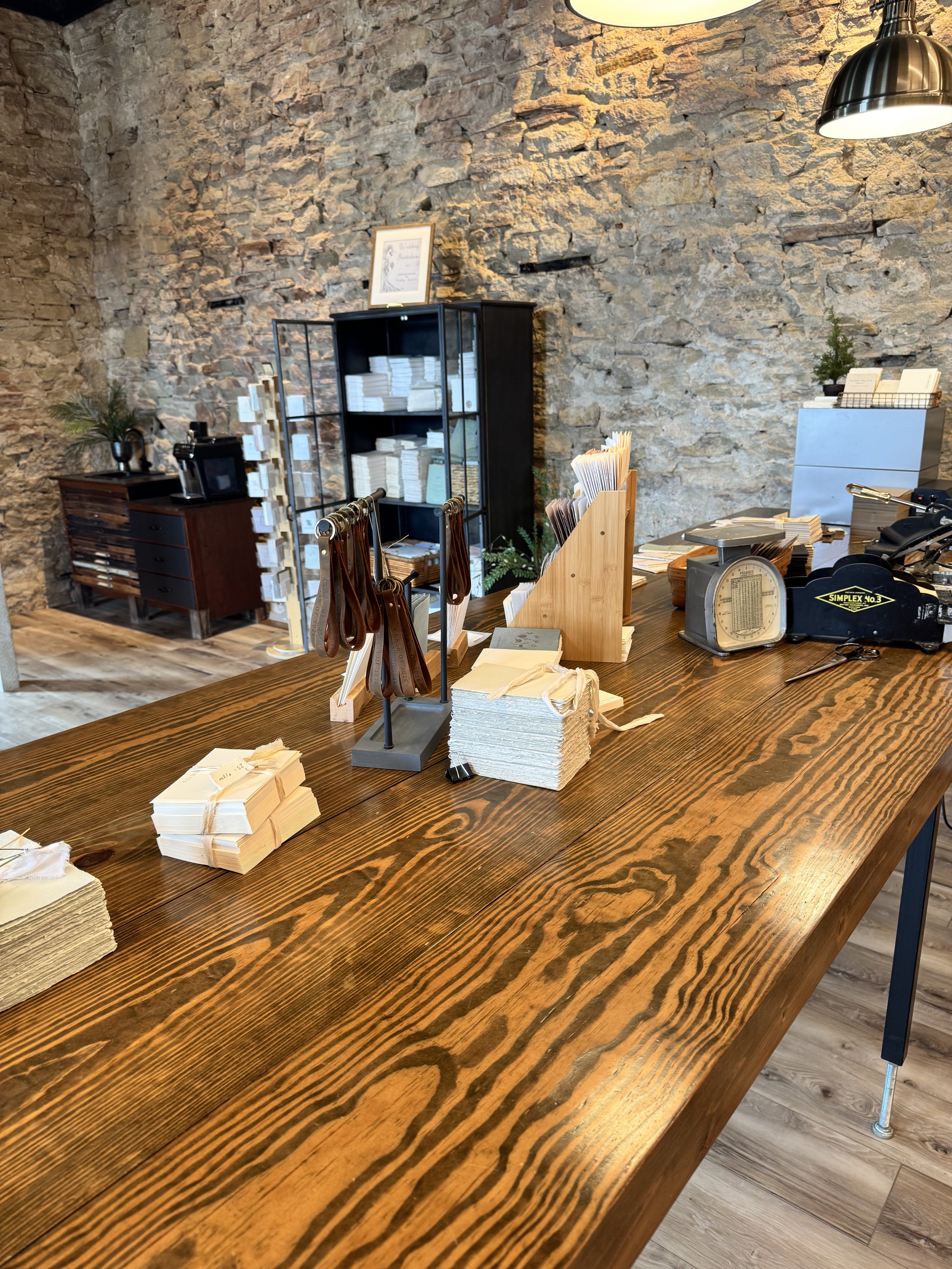 Interior of a retail or office space with a large wooden table holding fabric swatches and samples, against a rustic stone wall, with shelving and scales in the background.
