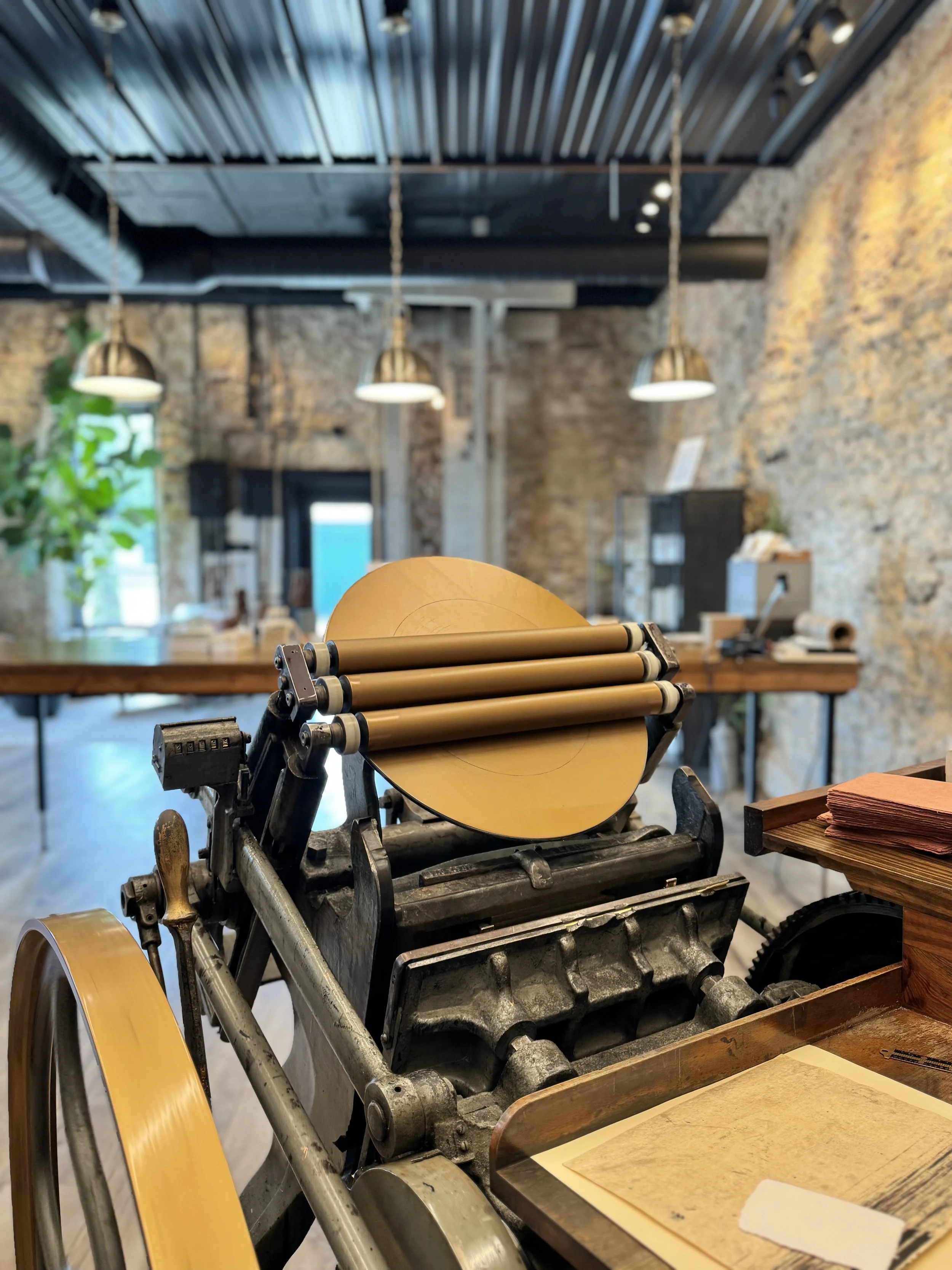 A vintage printing press with rollers and a wooden handle, placed on a table in a rustic, brick-walled room with exposed metal ceiling and hanging pendant lights.